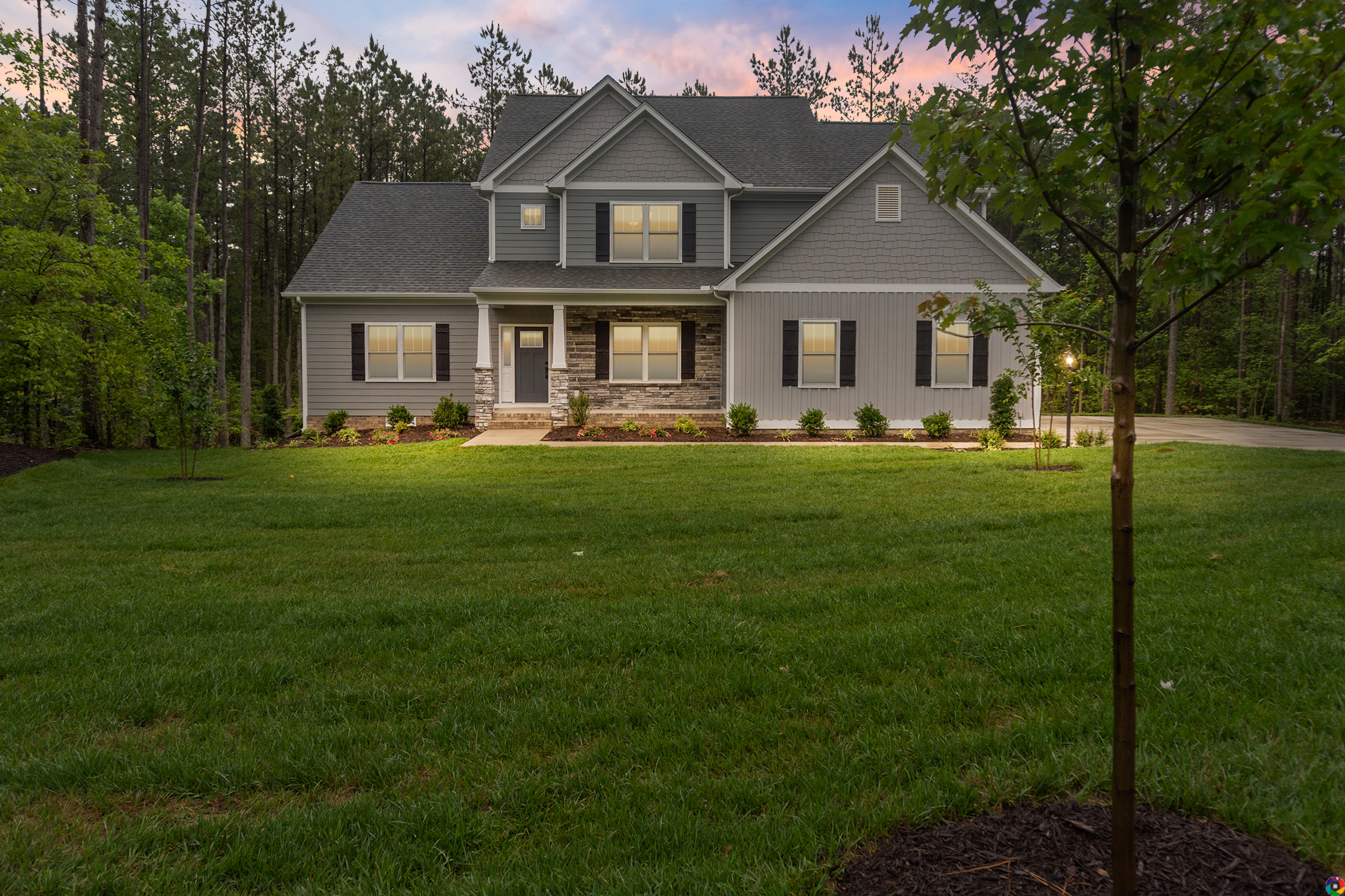 A two-story house with gray siding and a stone facade is set against a backdrop of tall trees, with a well-manicured lawn and lit windows during dusk.