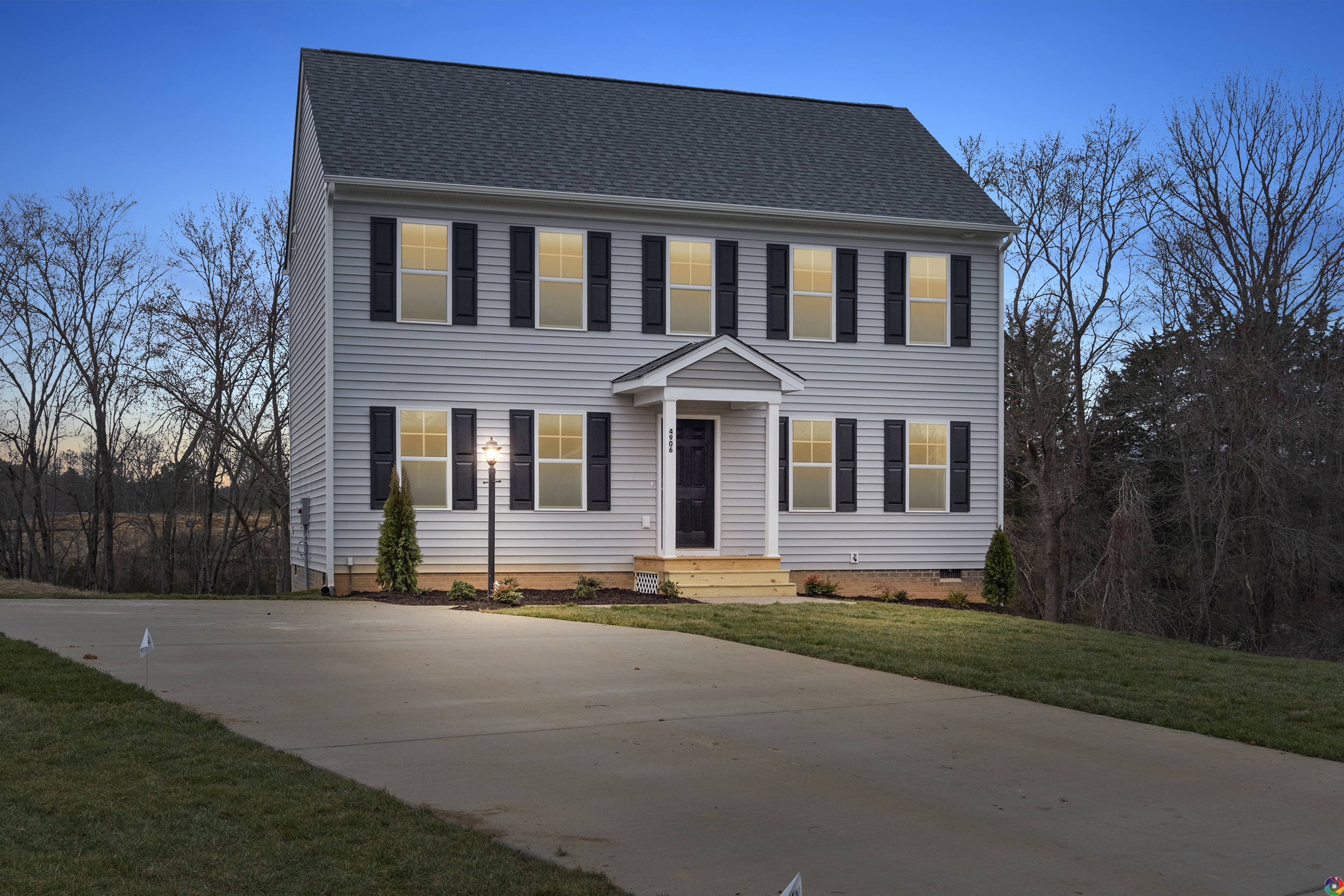 A two-story suburban house with white siding and black shutters, featuring a front porch and a lamp post. The house is lit from within and the surrounding lawn and driveway are visible at dusk.