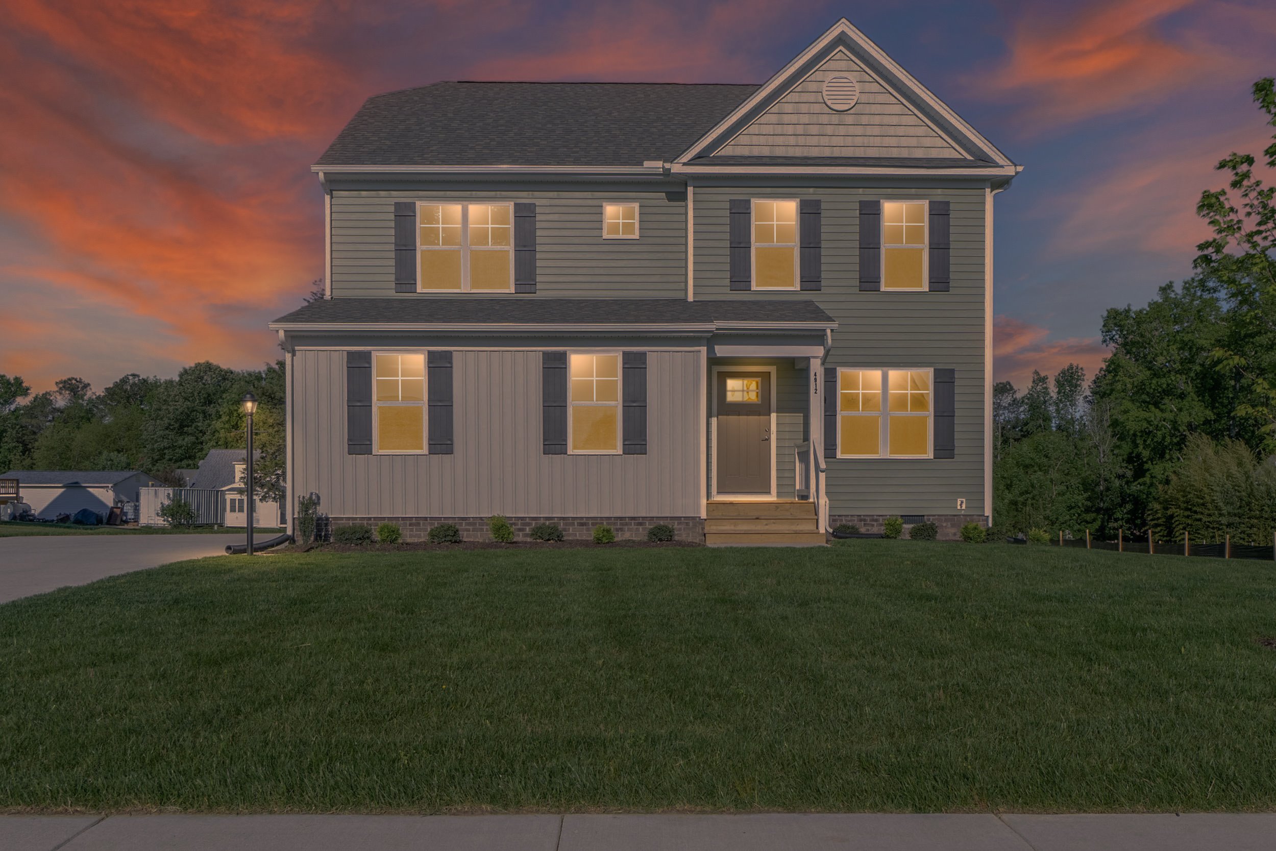Two-story house with illuminated windows, gray siding, and a green lawn in front, set against a dusk sky with orange and purple hues.