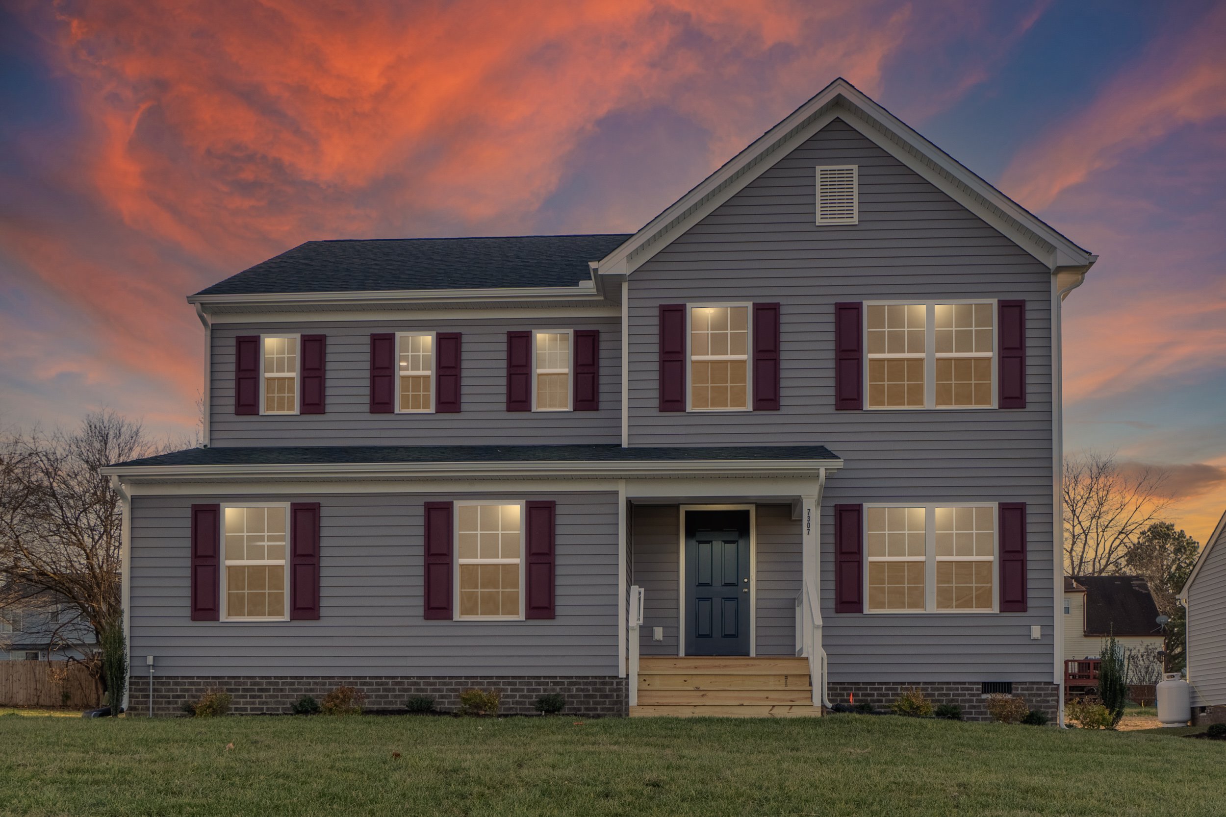 A two-story house with gray siding, a dark front door, and red shutters, lights on inside, stands before a dramatic sunset with orange and purple clouds in the sky.