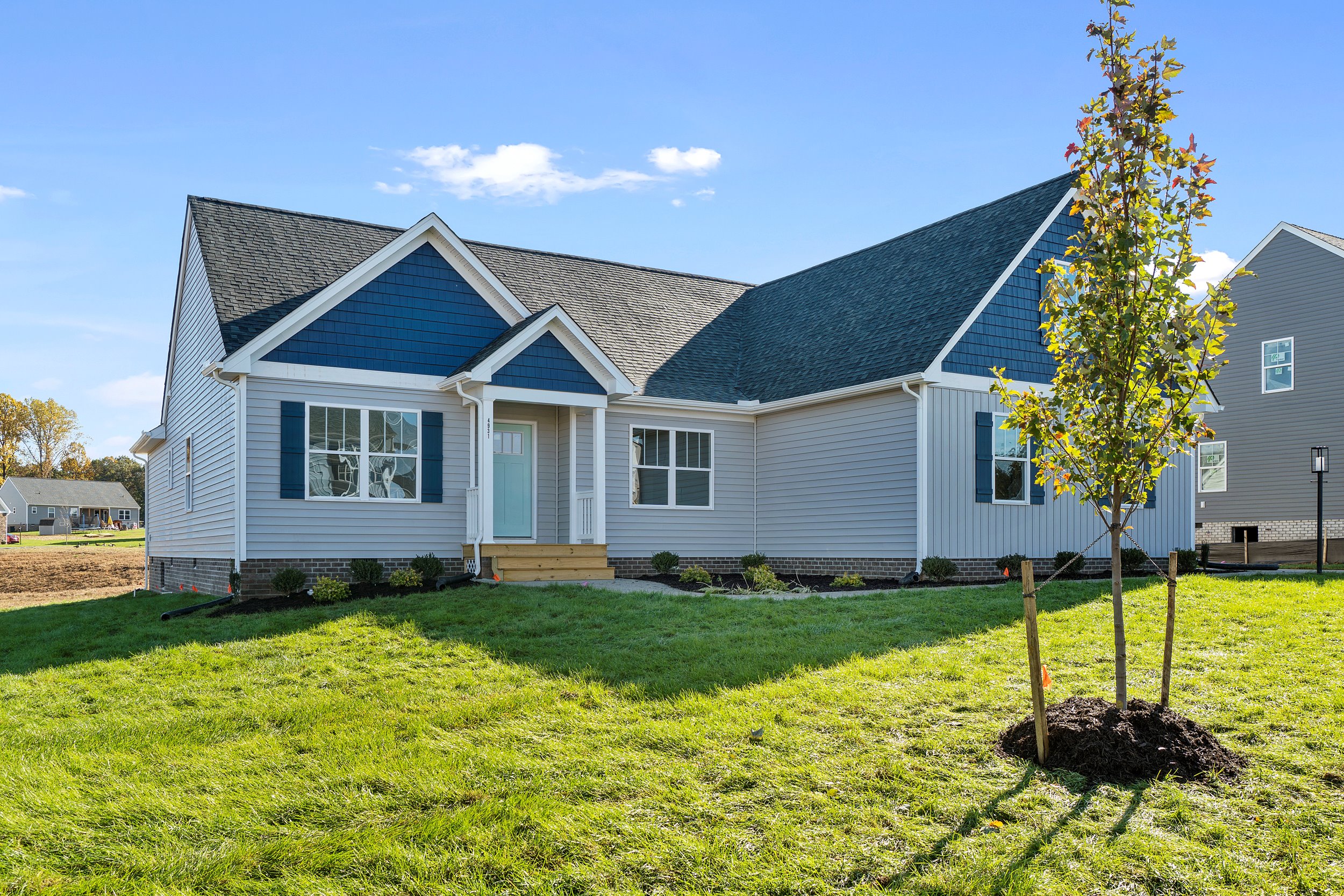 New suburban house with gray siding and dark blue shutters under a clear blue sky, featuring a neatly landscaped front yard and a newly planted tree.