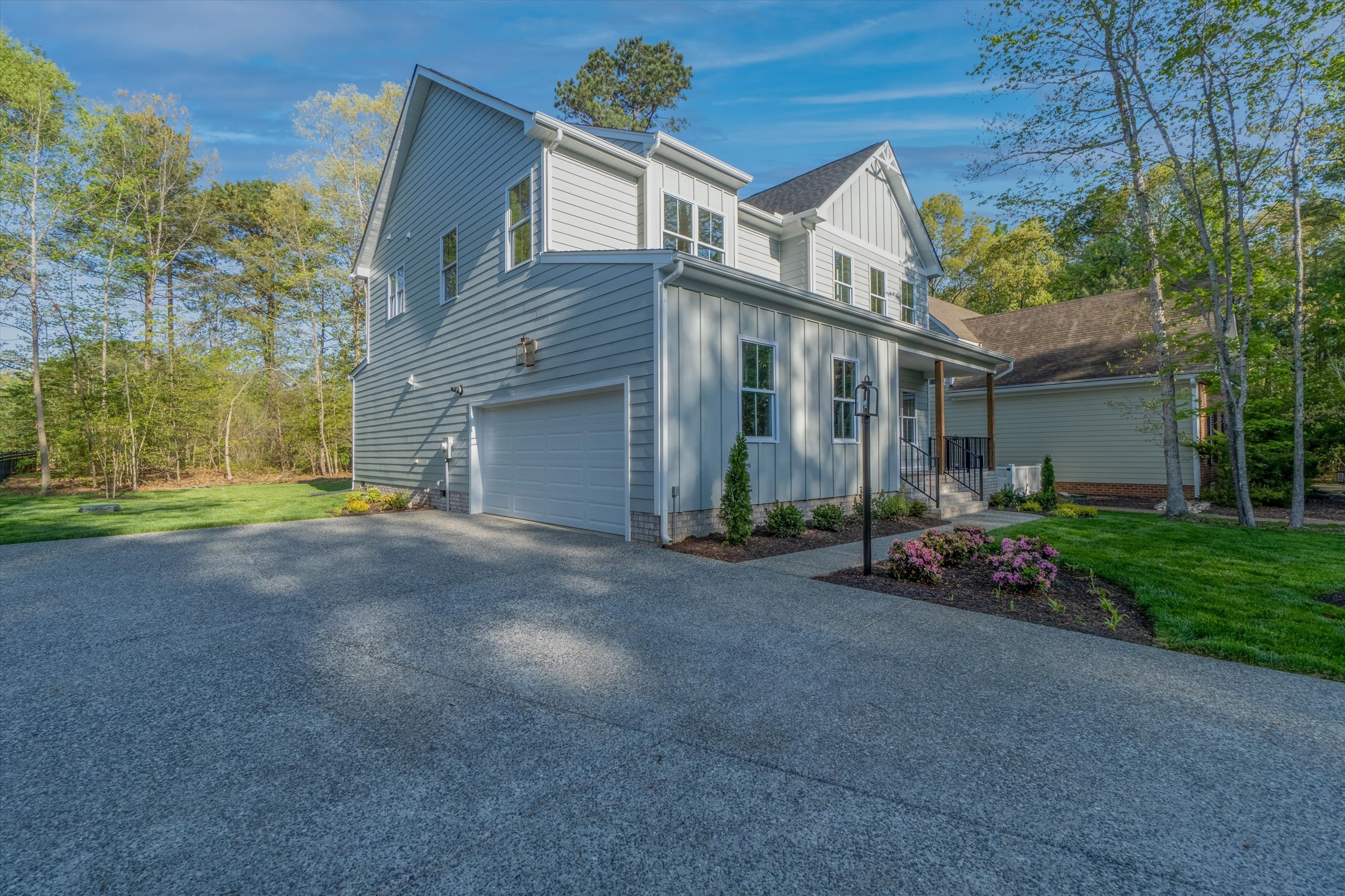 A two-story modern house with a grey exterior and attached garage, surrounded by a landscaped yard with trees and flowers, on a sunny day.