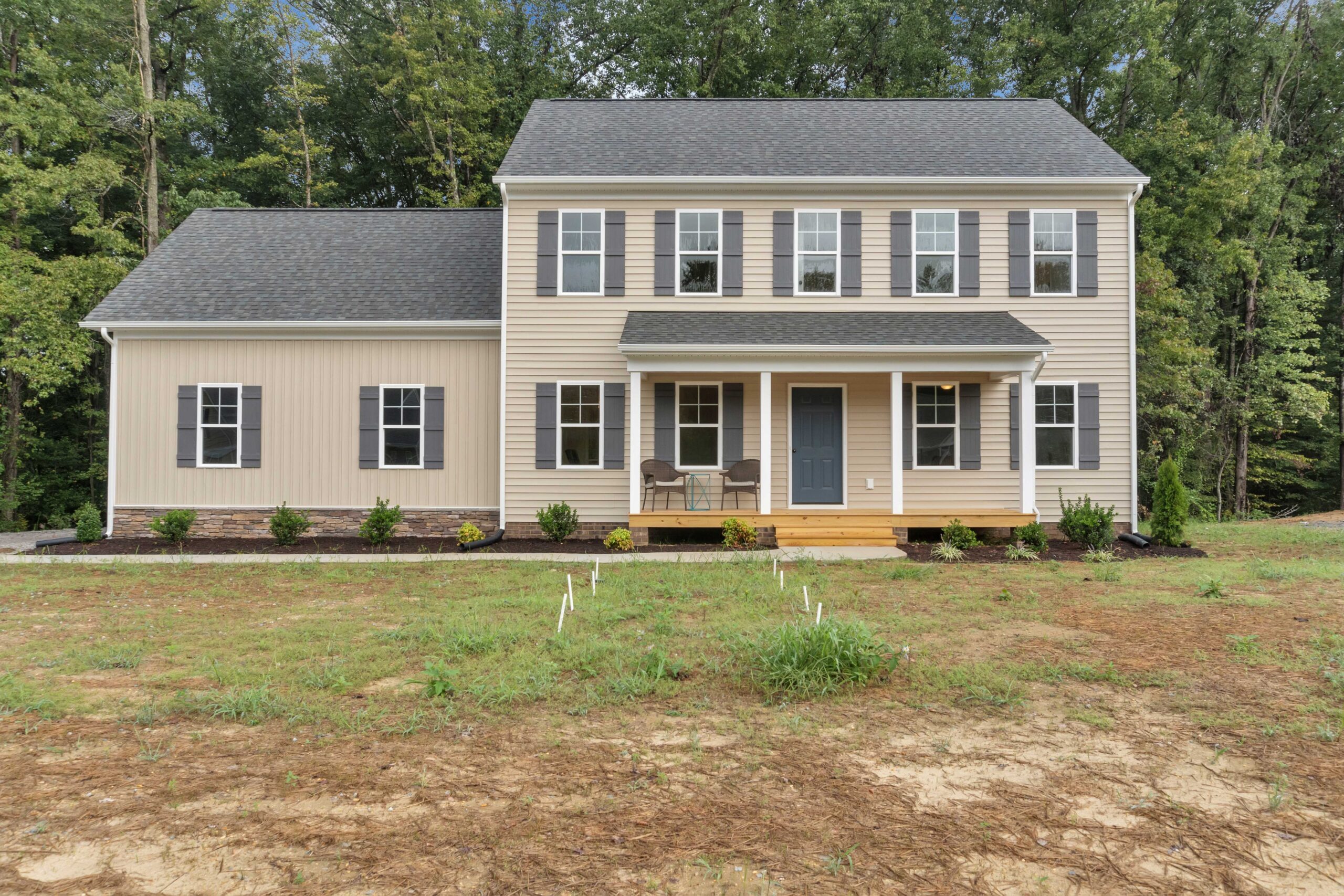 Two-story beige house with a dark gray roof, white trim, and a blue front door. There are six windows on the second floor and five on the first. The house is surrounded by a small, undeveloped yard.