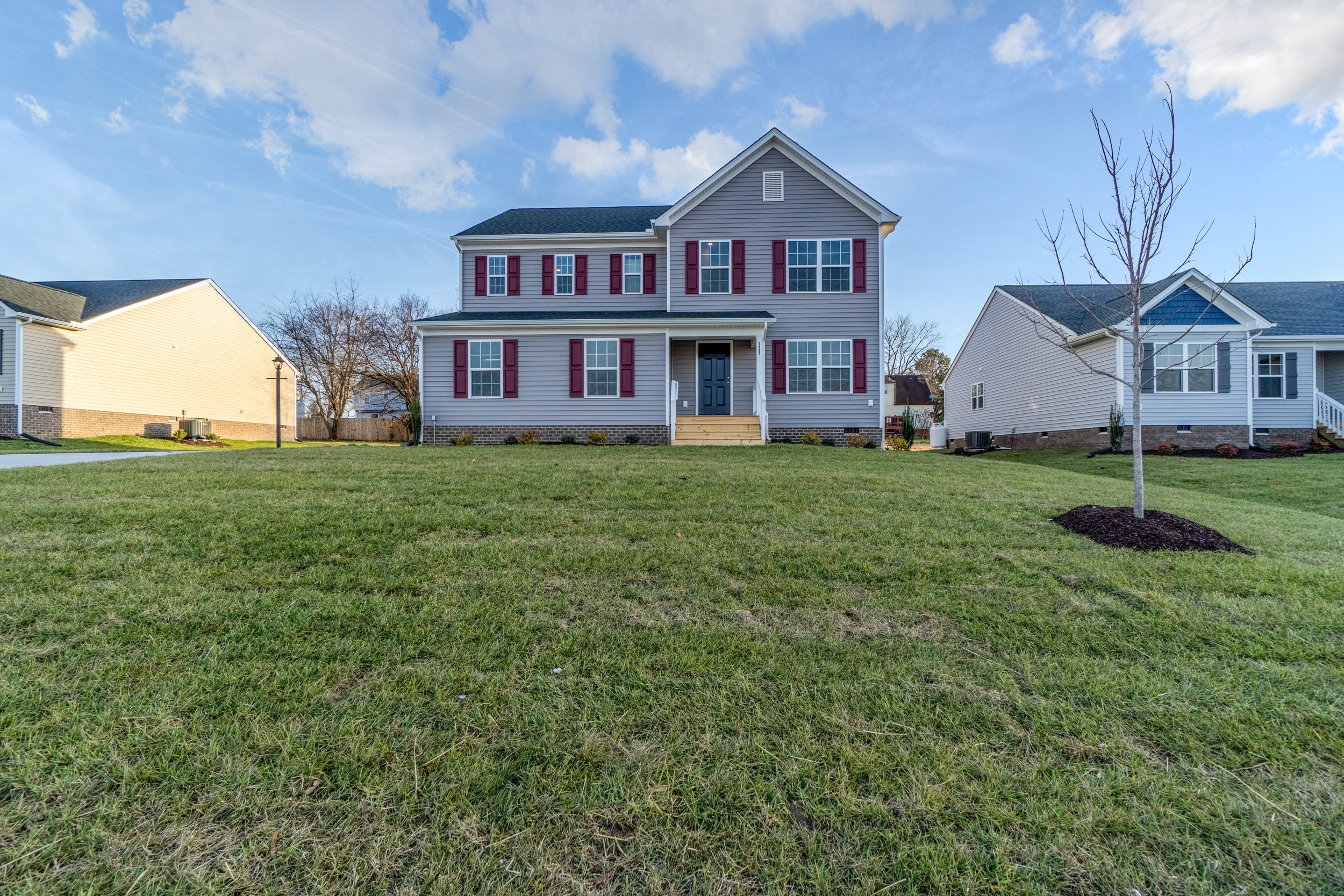 A two-story house with a grey exterior, red shutters, and a front porch, located on a grassy lawn with other similar houses nearby. The sky is partly cloudy.
