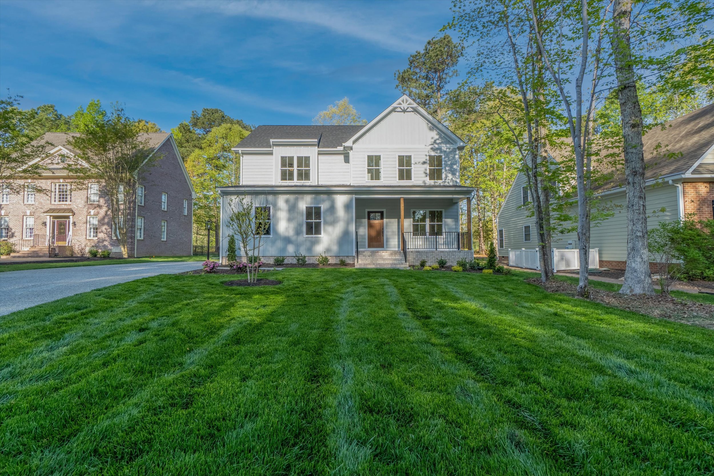 A two-story house with a front porch stands in the center, surrounded by a well-manicured lawn and trees. Another house is visible on the left side, with clear skies and ample daylight illuminating the scene.