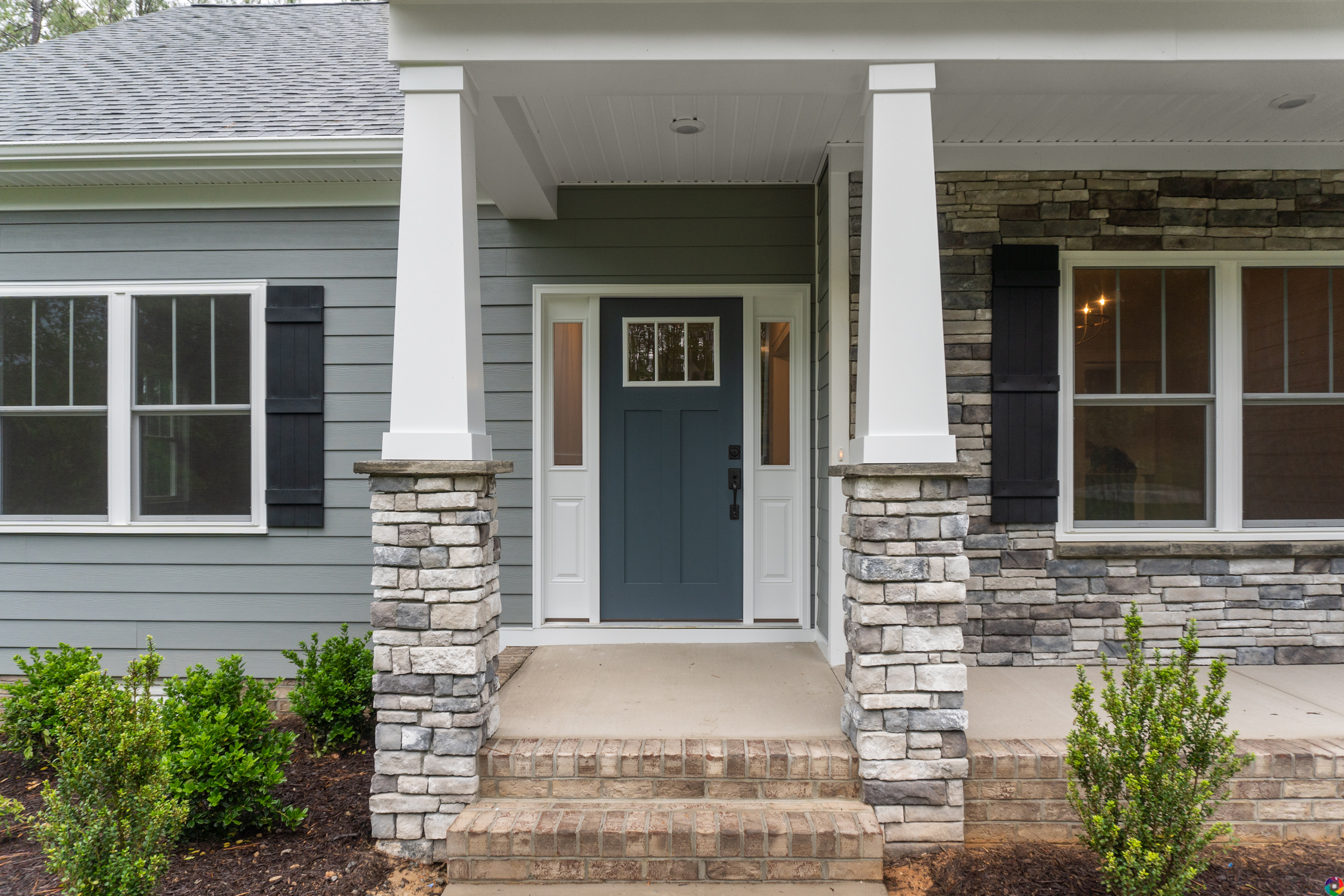 Front porch of a house with stone columns, a blue front door with sidelights, and gray siding. The porch has neatly landscaped bushes in front.