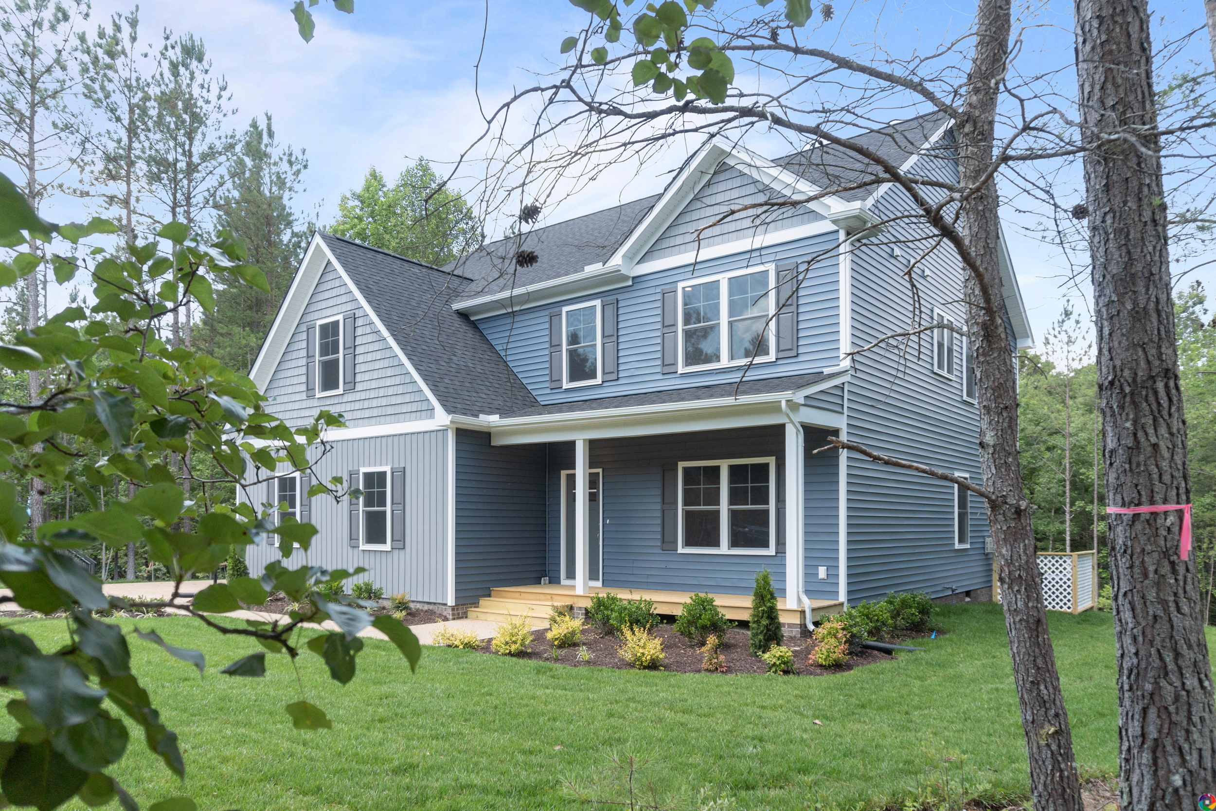 A two-story blue house with white trim and a covered porch, surrounded by a green lawn and trees, under a clear sky.