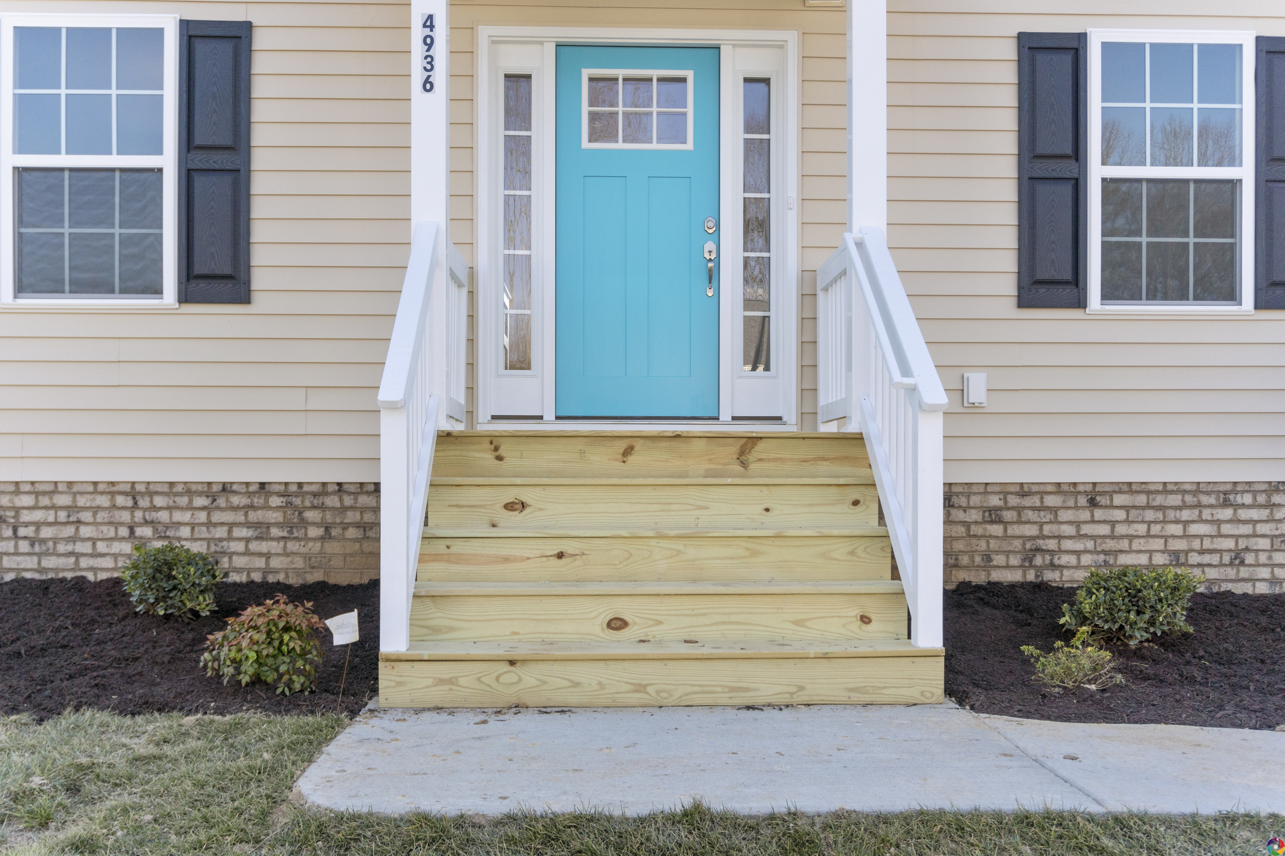 Front entrance of a house with a turquoise door, beige siding, white trim, and black shutters, featuring wooden steps with white railings and a small garden area on either side.