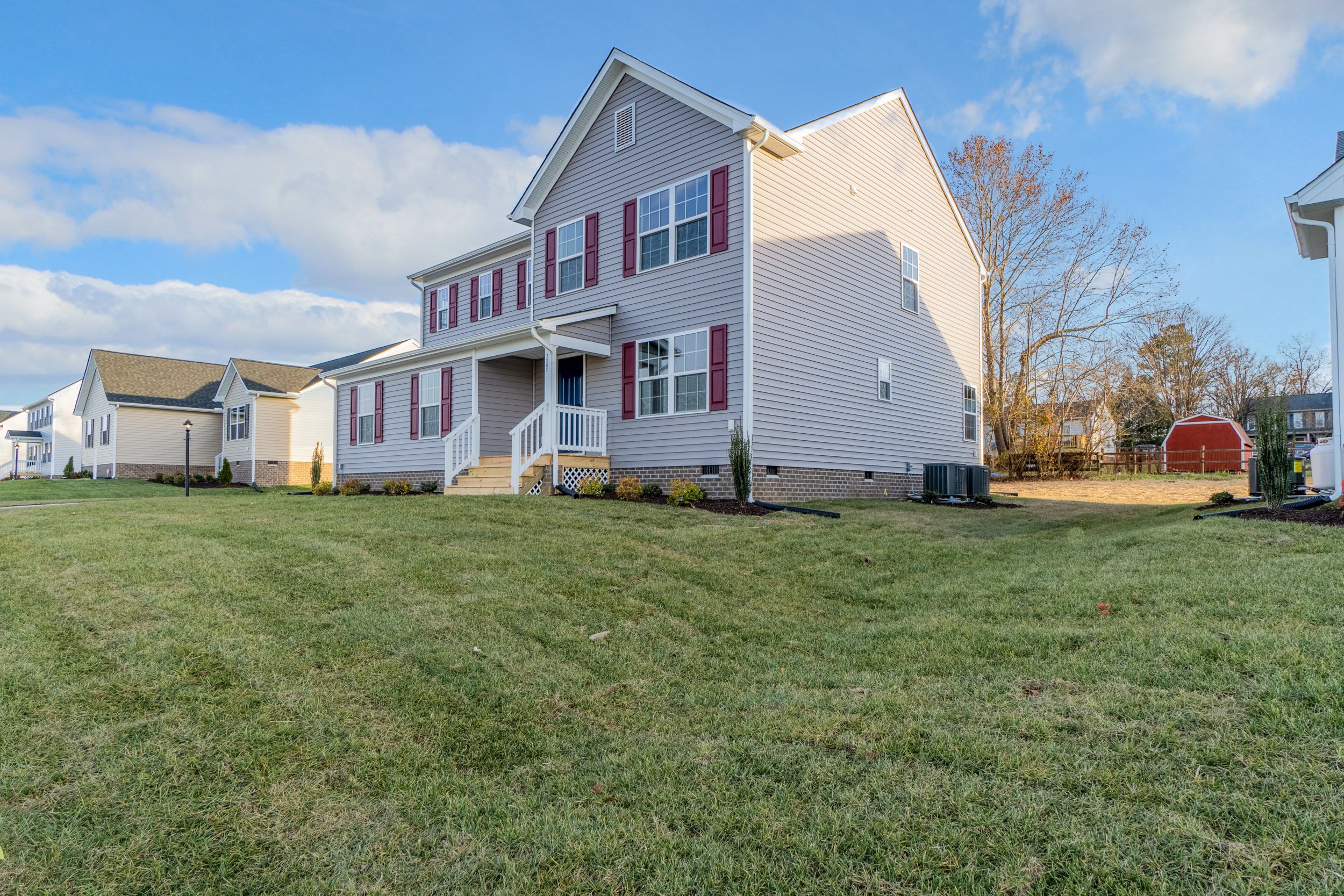 A two-story white house with red shutters stands on a large grassy lawn under a blue sky with some clouds.