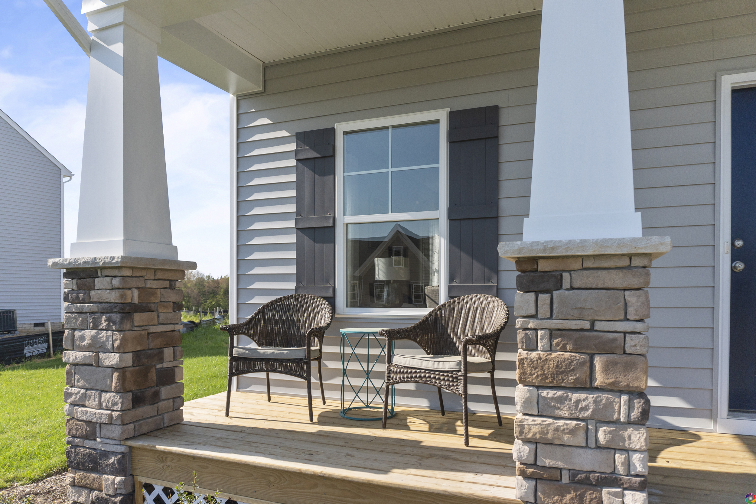 Two wicker chairs and a small table on a wooden porch with stone columns, facing a modern house with large windows.