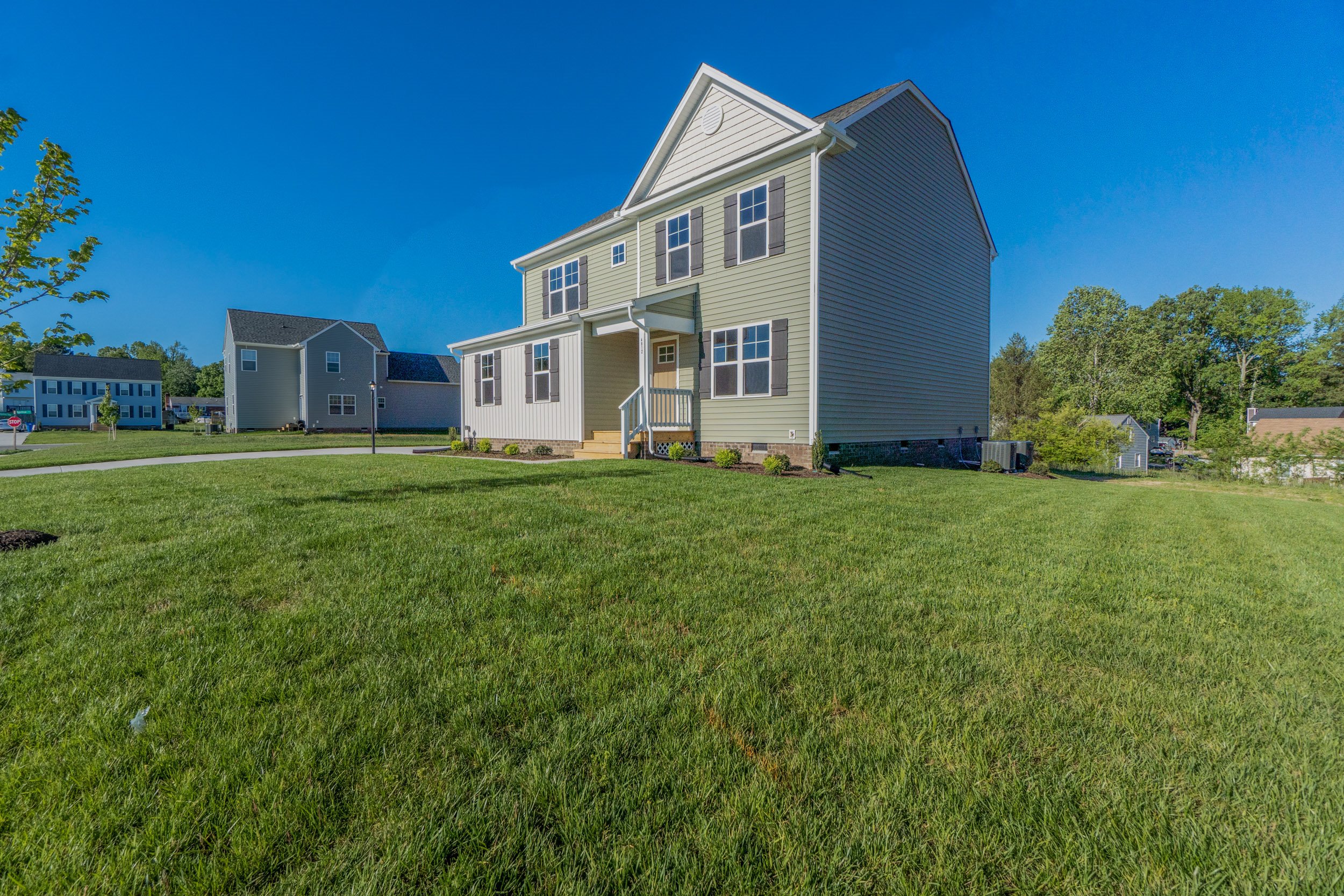 A beige two-story house with a front porch is situated on a large, well-maintained lawn under a clear blue sky. Other houses and trees are visible in the background.