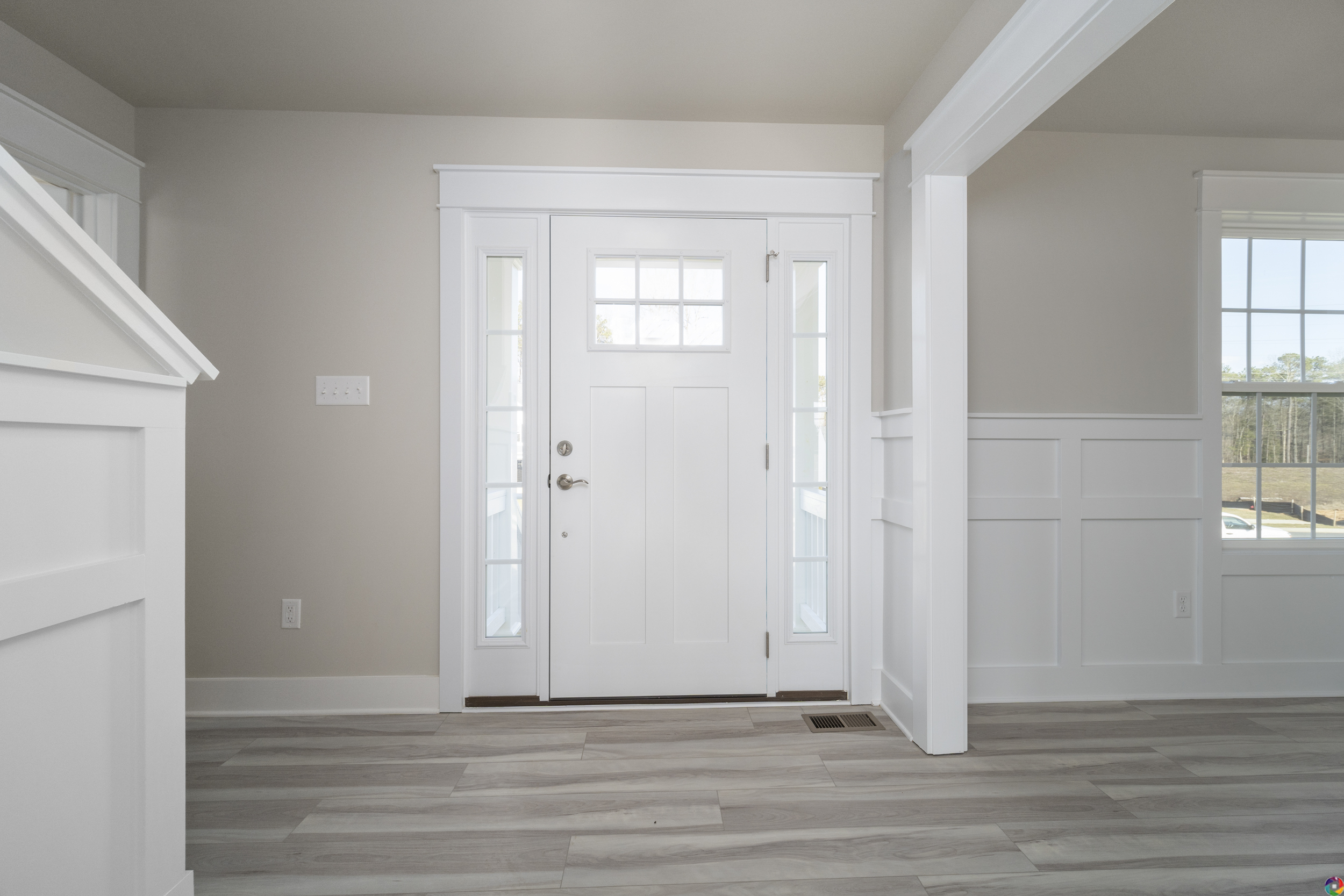 A white front door with glass panels, surrounded by sidelights and a transom window, opens into an interior with light hardwood floors and wainscoting.