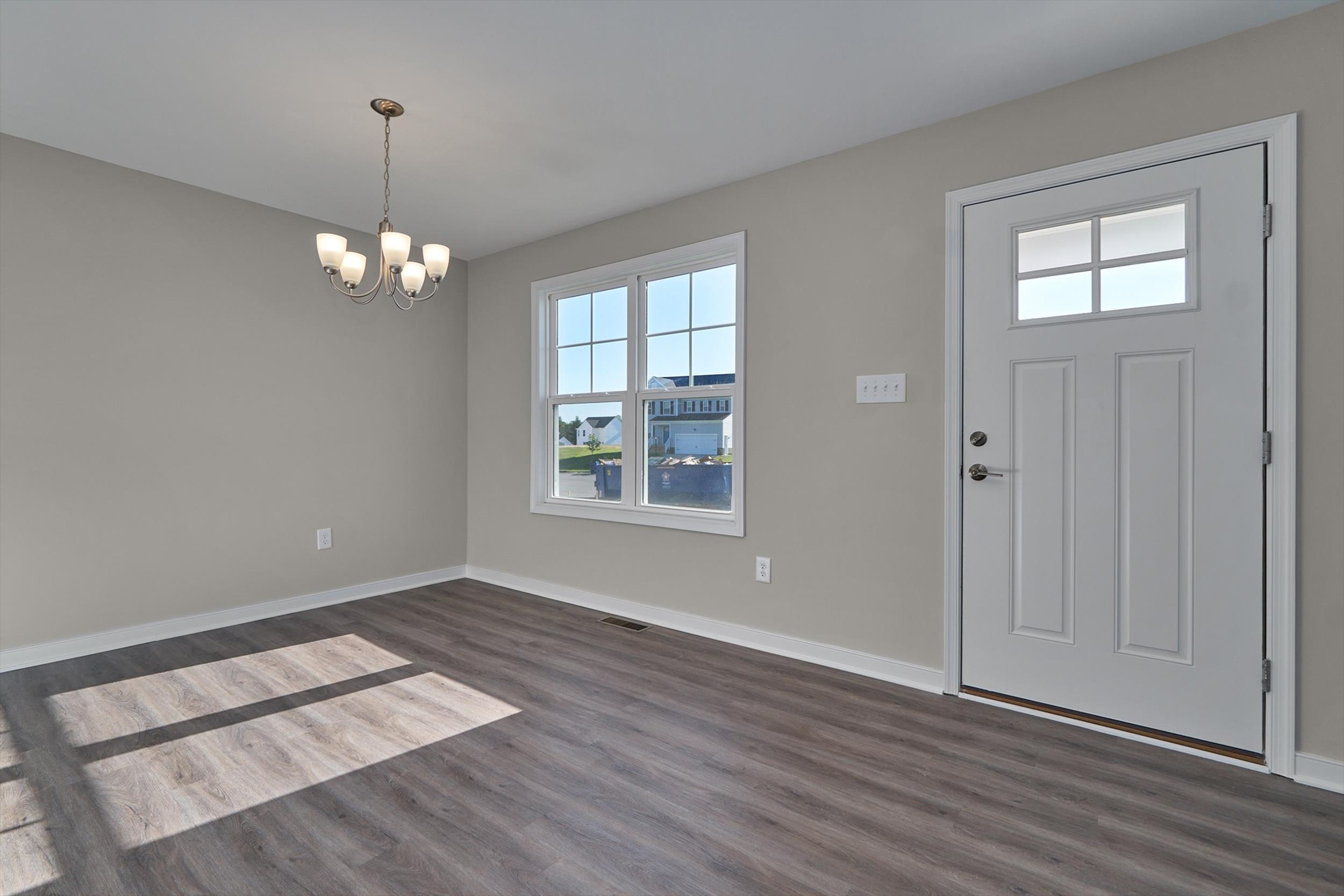 A bright, empty room with large windows, a chandelier, wooden flooring, and a white door with a small window.