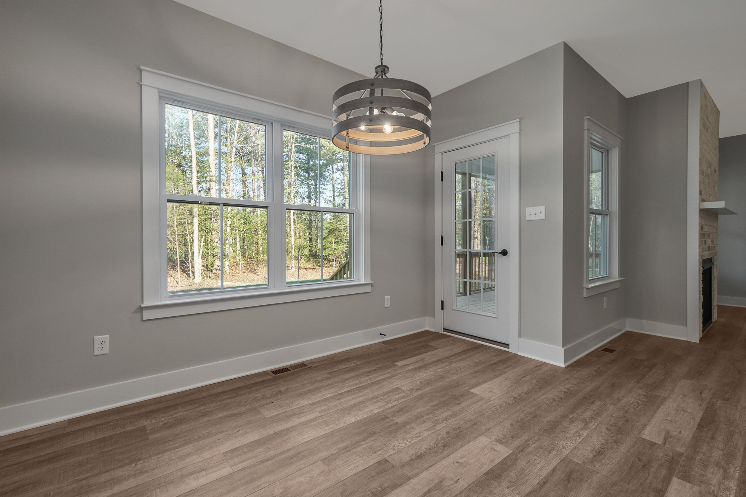 A room with light gray walls, large windows, a glass door, hardwood floors, and a modern light fixture. Outside, trees are visible through the windows.