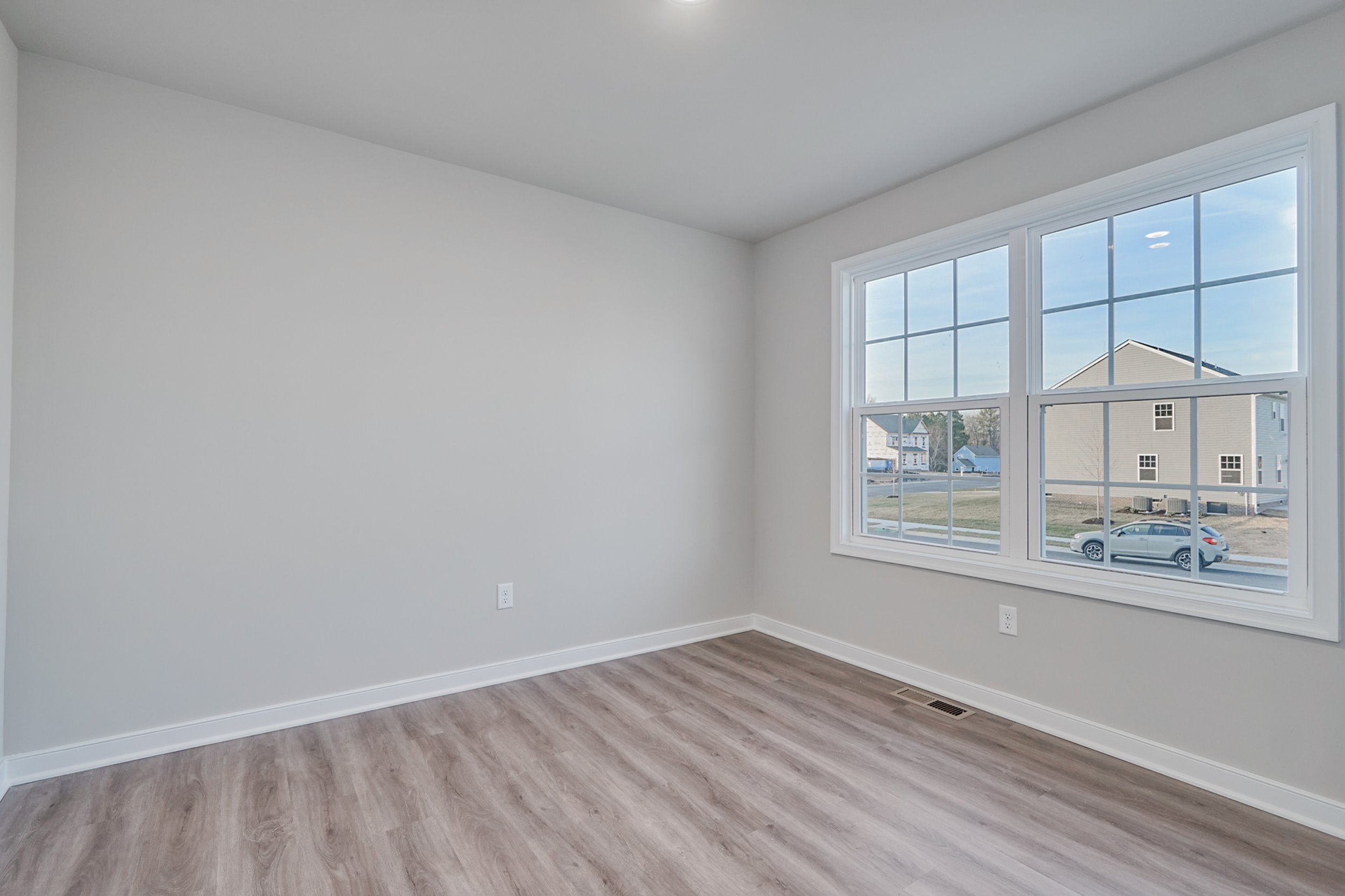 A small empty room with light-colored walls and wood flooring. A large window on one wall provides a view of a suburban neighborhood.