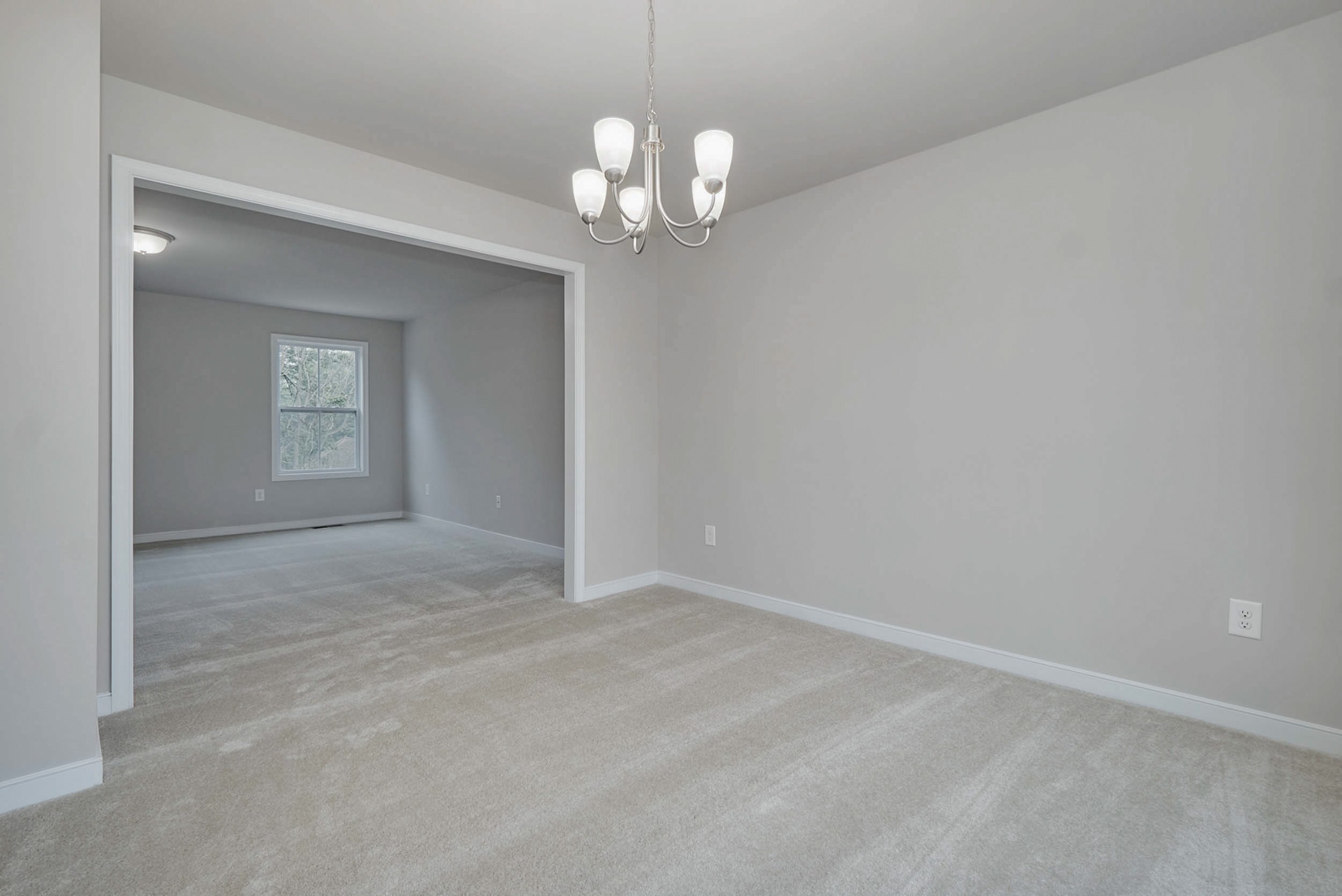 Empty room with beige carpeted flooring, a white ceiling, and light gray walls. A chandelier hangs from the ceiling. Another room with a window is visible through the open doorway.