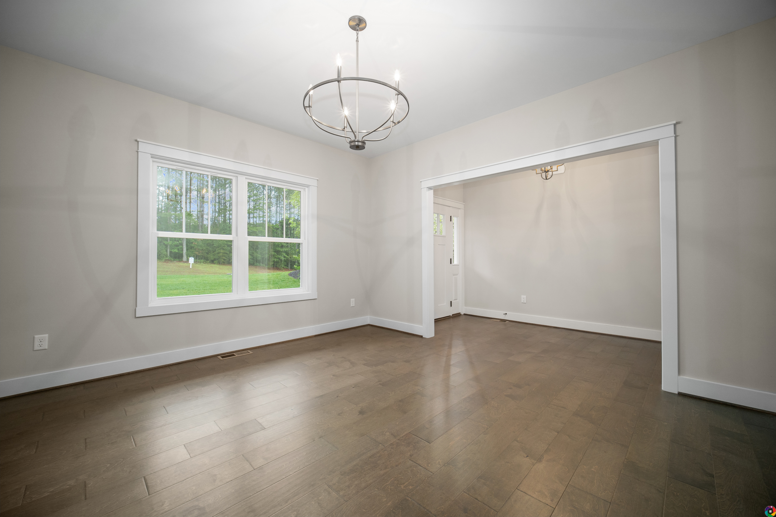 An empty room with light gray walls, hardwood floors, large window, and modern light fixtures. Doorway leads to an adjoining room.