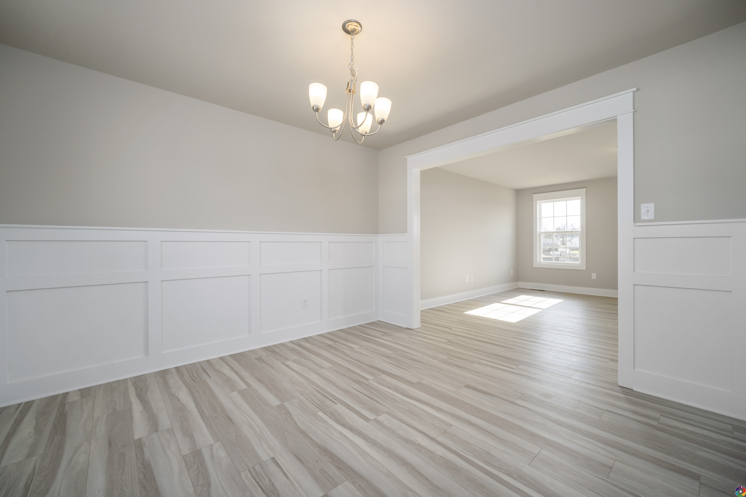 A bright, empty room with light wood floors, white wainscoting, and a chandelier. An open doorway leads to another sunlit room with a large window.