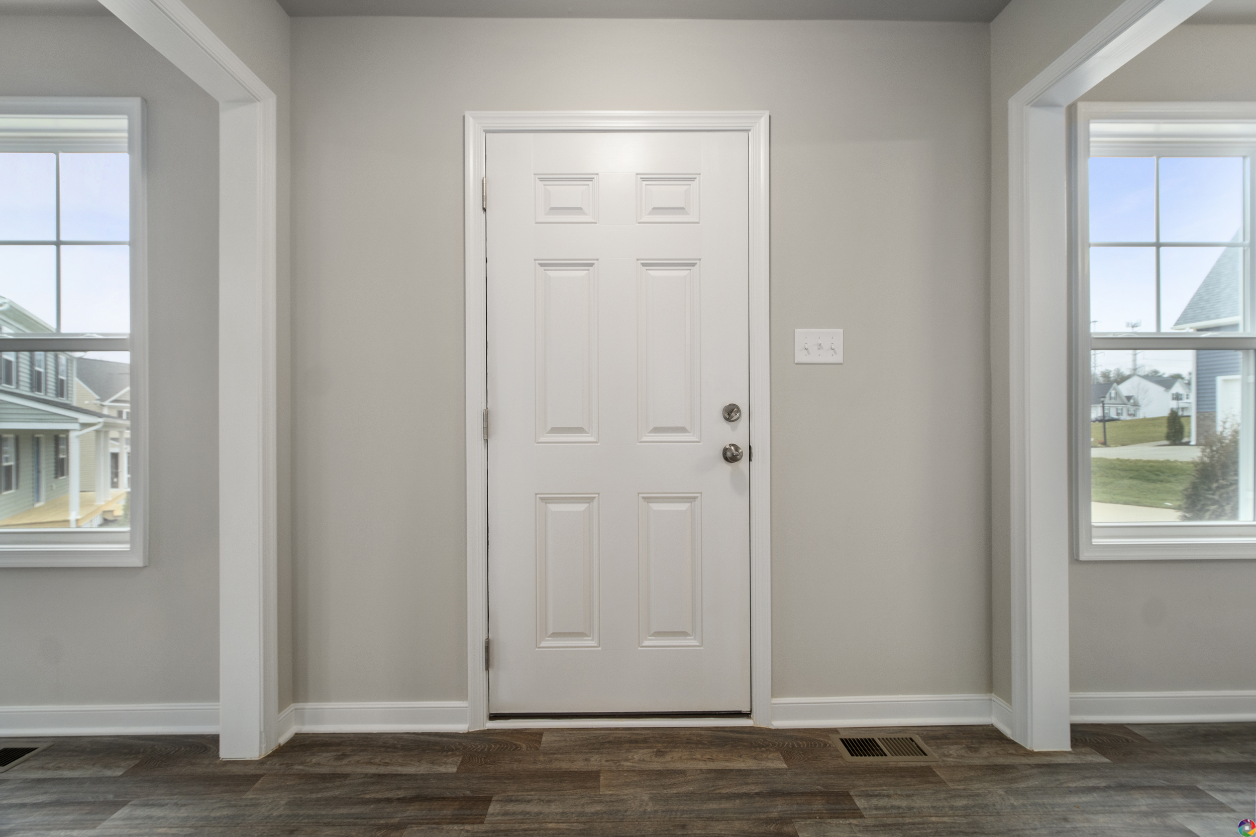 Interior view of a house entrance featuring a white door centered between two windows, with wood flooring and neutral-colored walls.