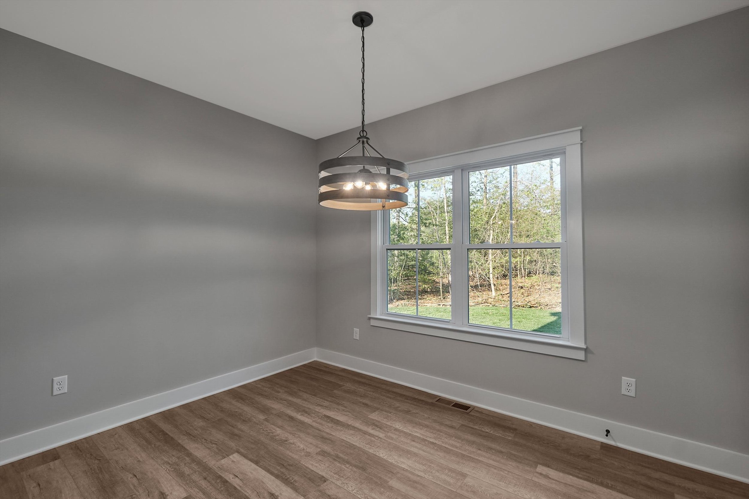 A vacant room with gray walls, a large window with a view of trees, a modern hanging light fixture, and wooden flooring.