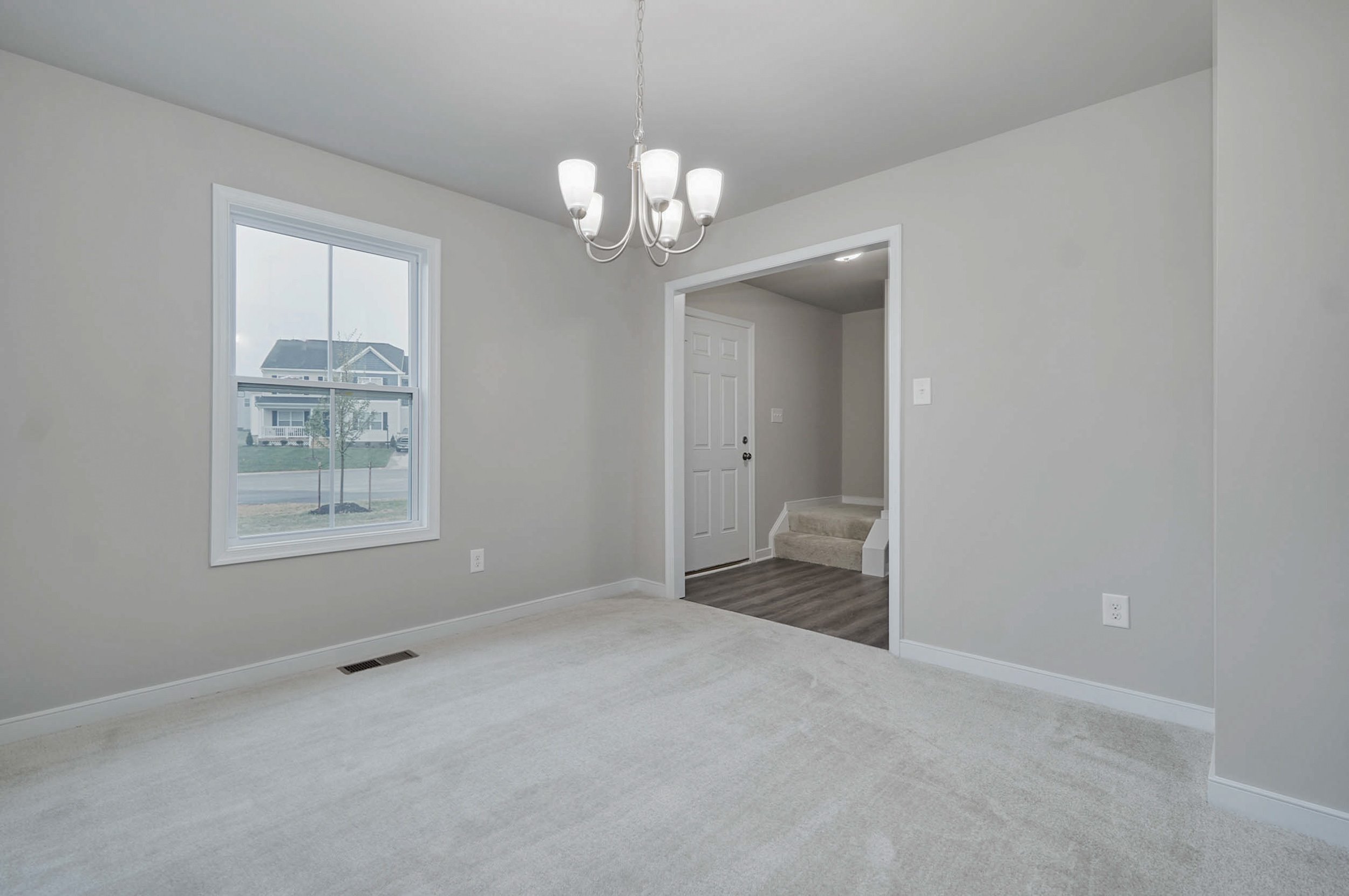 A simple, empty room with light gray walls, a white ceiling, and beige carpet. The room has a window, a chandelier, and a doorway leading to another room with wooden flooring.