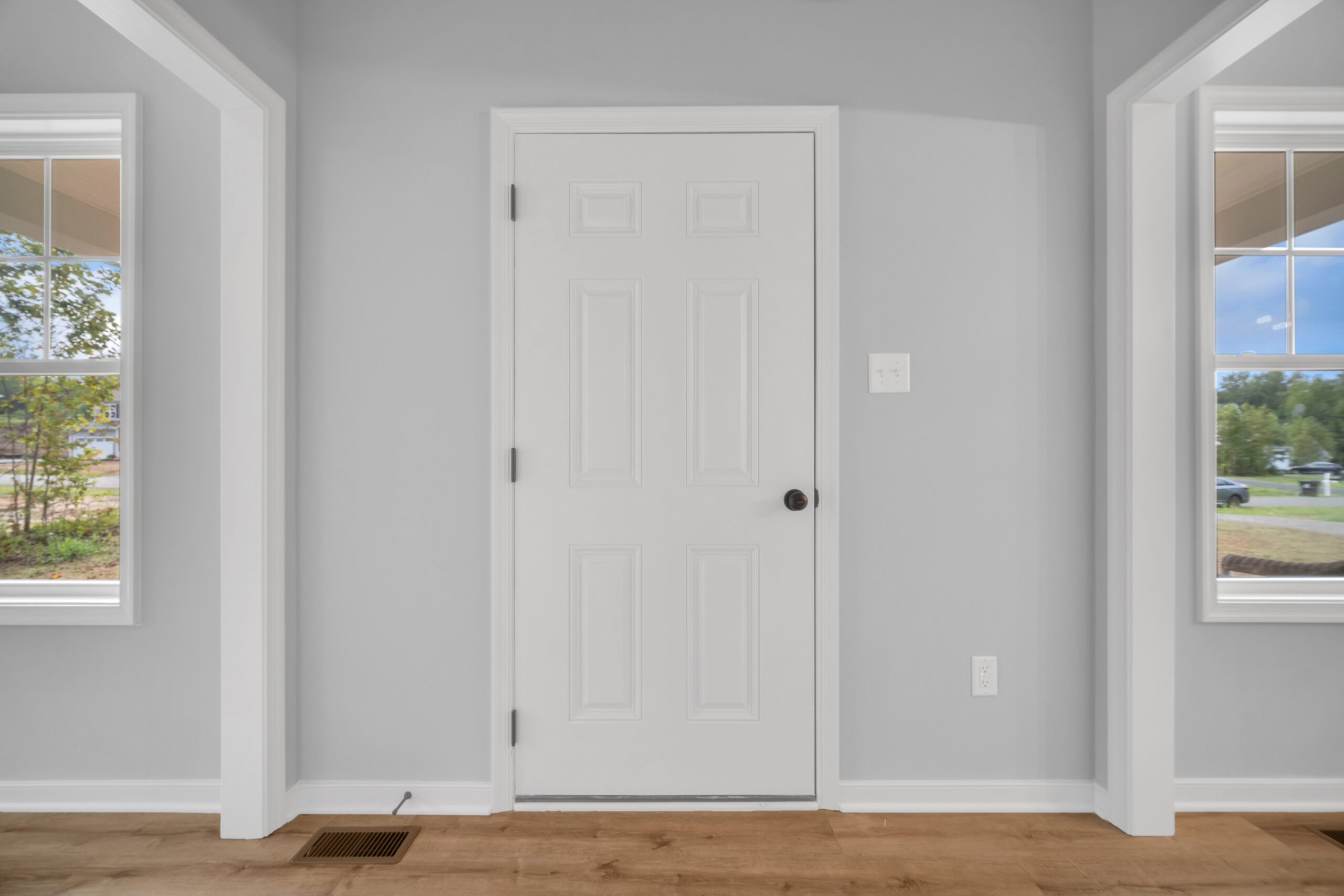 A white closed door in the center of a light gray wall, flanked by two windows showing an outdoor view, with wooden flooring beneath.