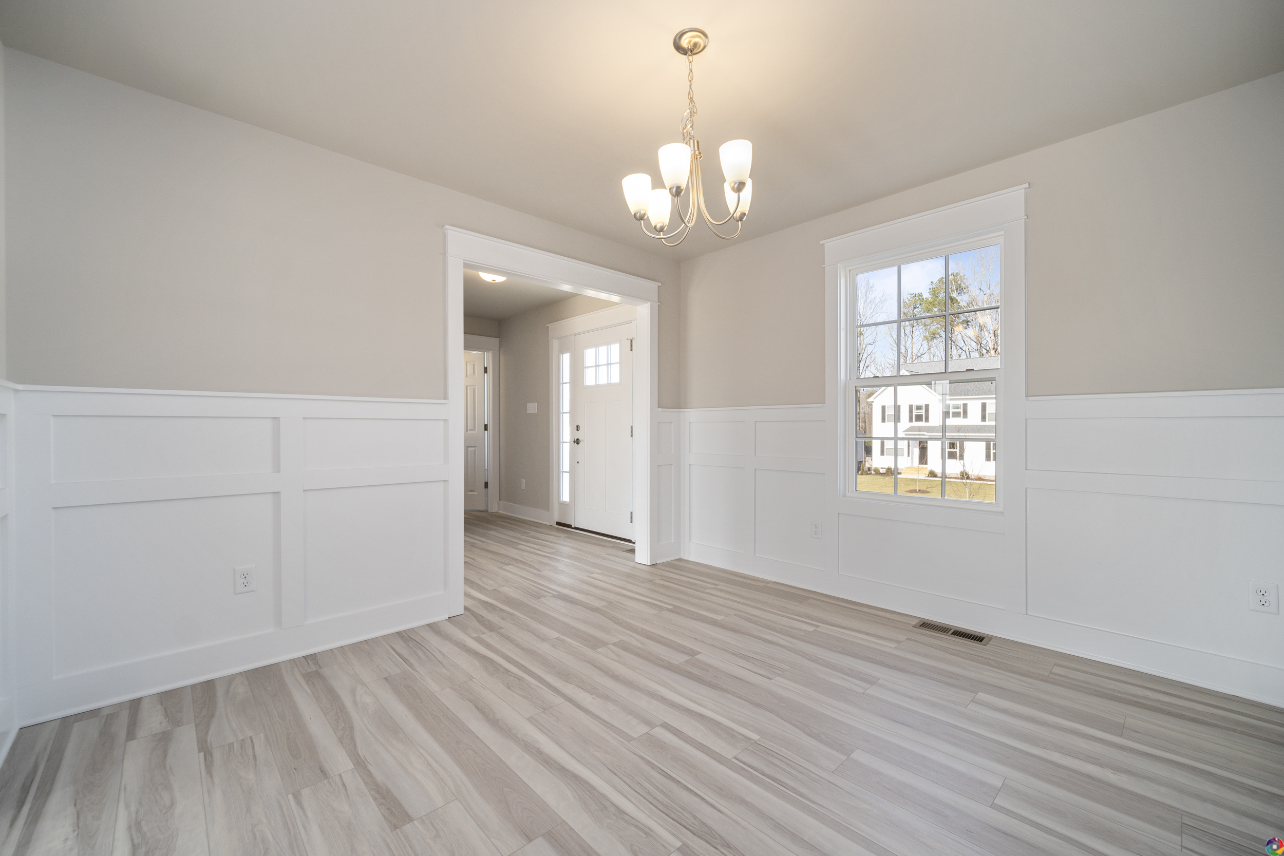 A bright, empty room with light gray wood flooring, white wainscoting walls, a window, and a chandelier. An open doorway leads to a hallway with a front door and additional windows.