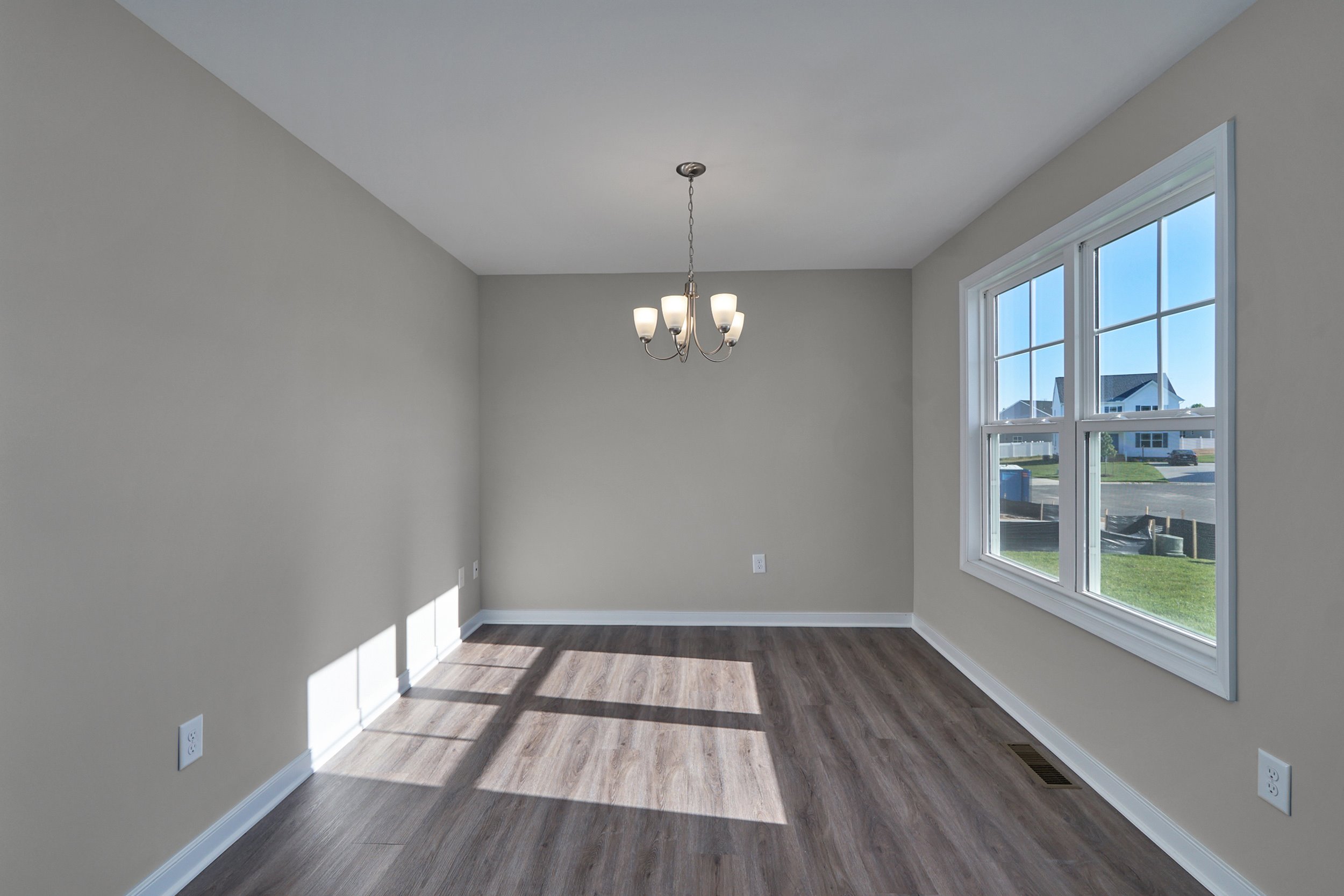 An empty room with light gray walls, wood flooring, a central ceiling light fixture, and a large window letting in natural light.