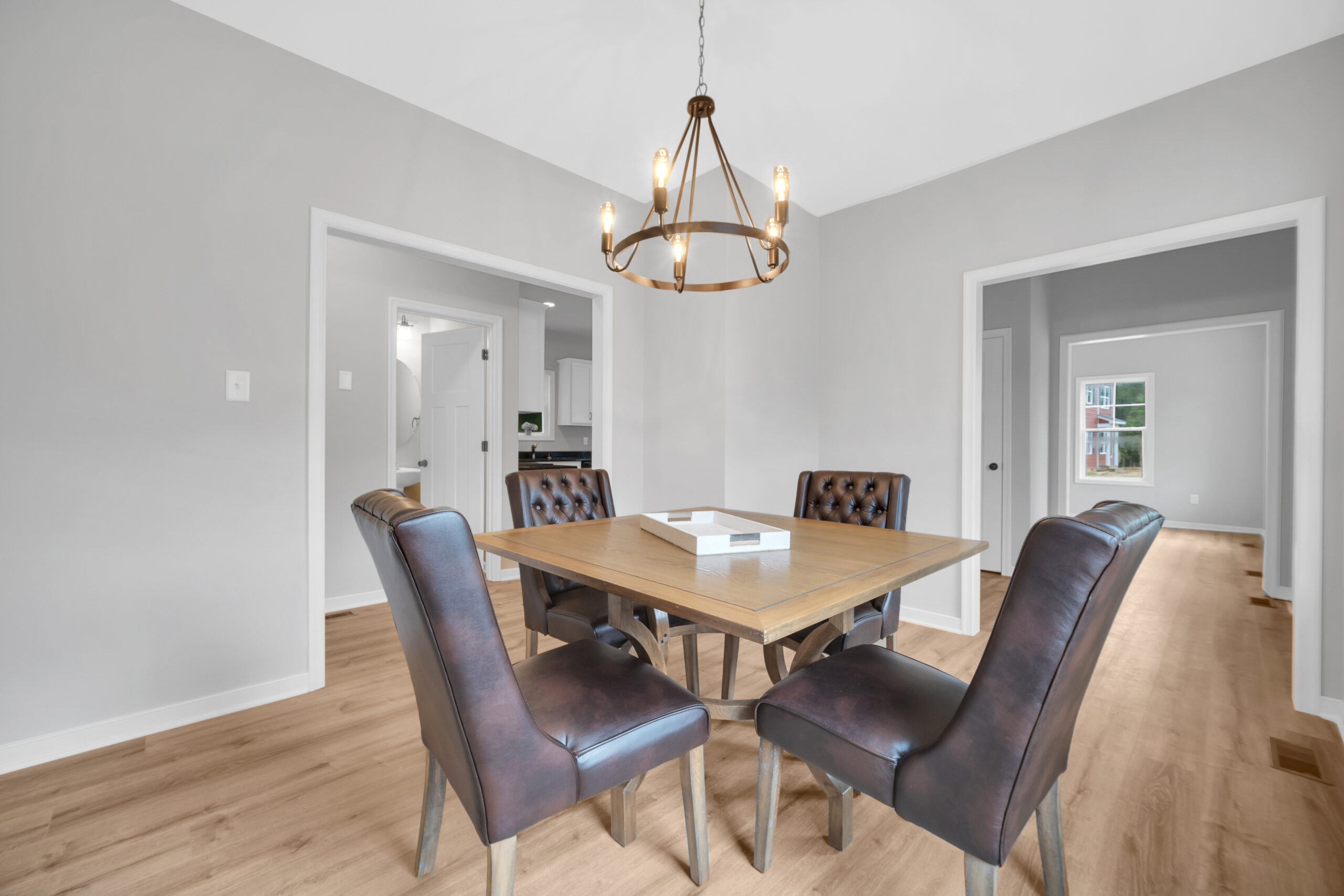 A dining room with a wooden table surrounded by four black padded chairs, a modern chandelier overhead, light wood flooring, and white walls with open doorways leading to adjacent rooms.
