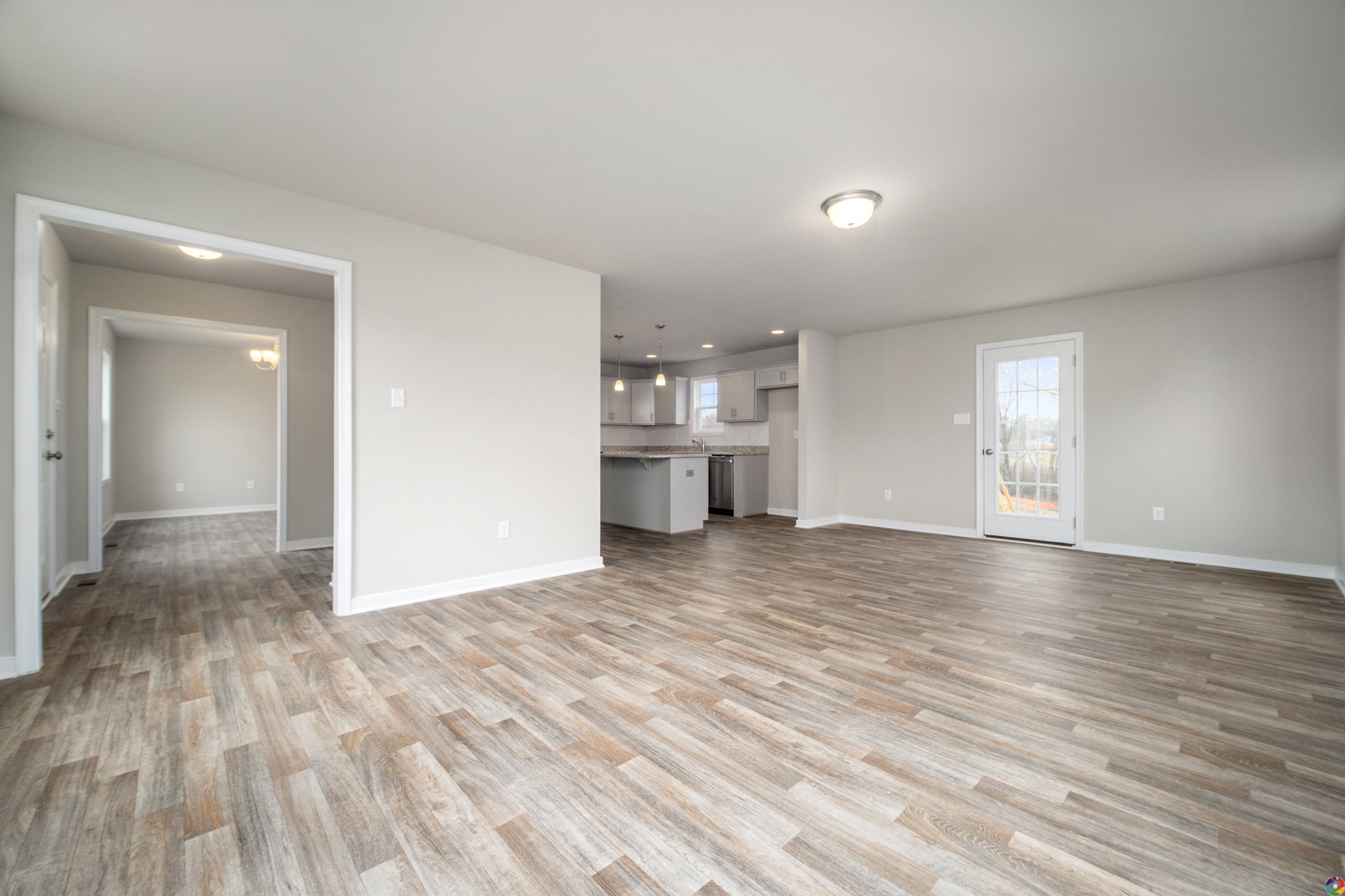 An empty room with wood-style flooring, light gray walls, a ceiling light, and a view of a connected kitchen and dining area. Doorways lead to additional rooms.