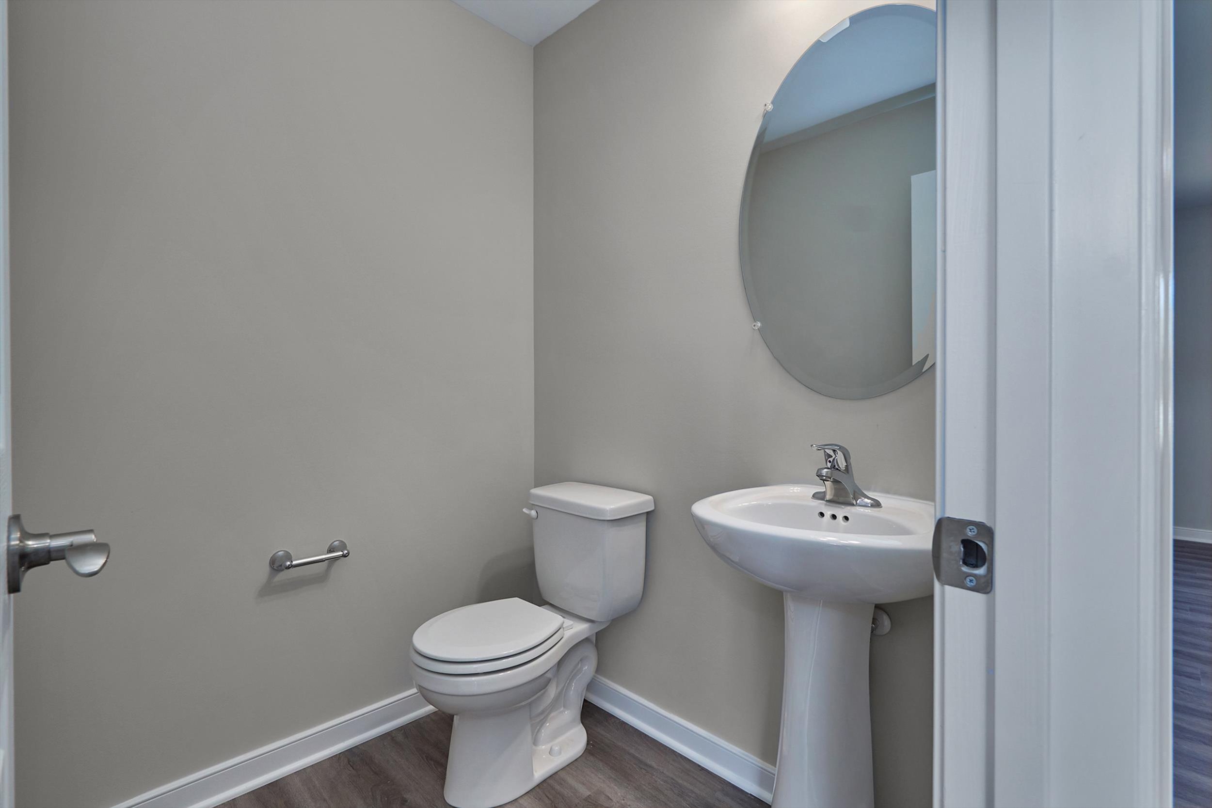 A small bathroom with a toilet, pedestal sink, and oval mirror on the wall. The floor has wood-like tiles, and the walls are painted light gray.