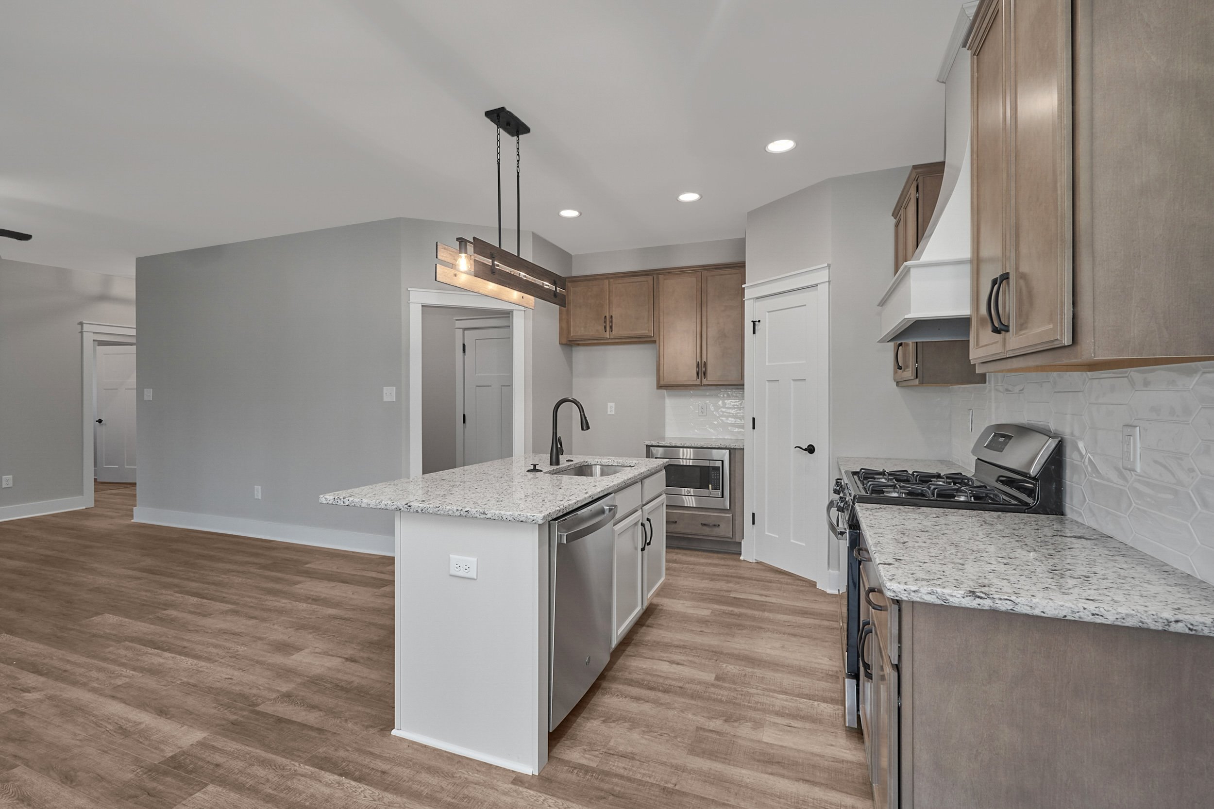 A modern kitchen with light wood cabinetry, granite countertops, an island with a sink, a gas stove, stainless steel appliances, and hardwood floors. The kitchen is well-lit with recessed and pendant lights.