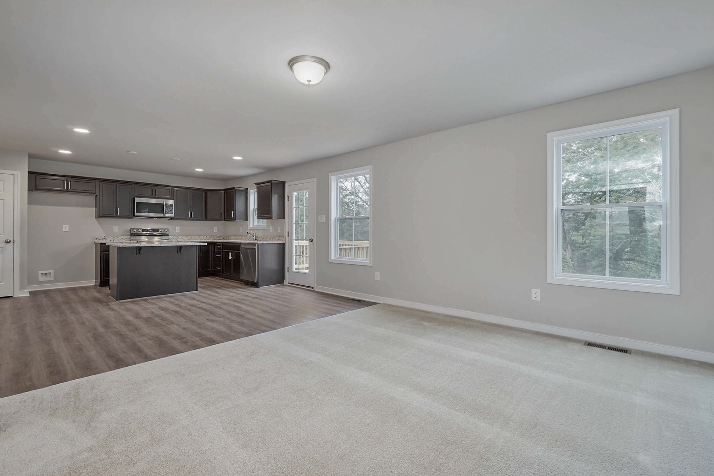An empty open-concept living area with a view of the kitchen. The room has light-colored walls, a mix of carpet and hardwood flooring, and large windows letting in natural light.