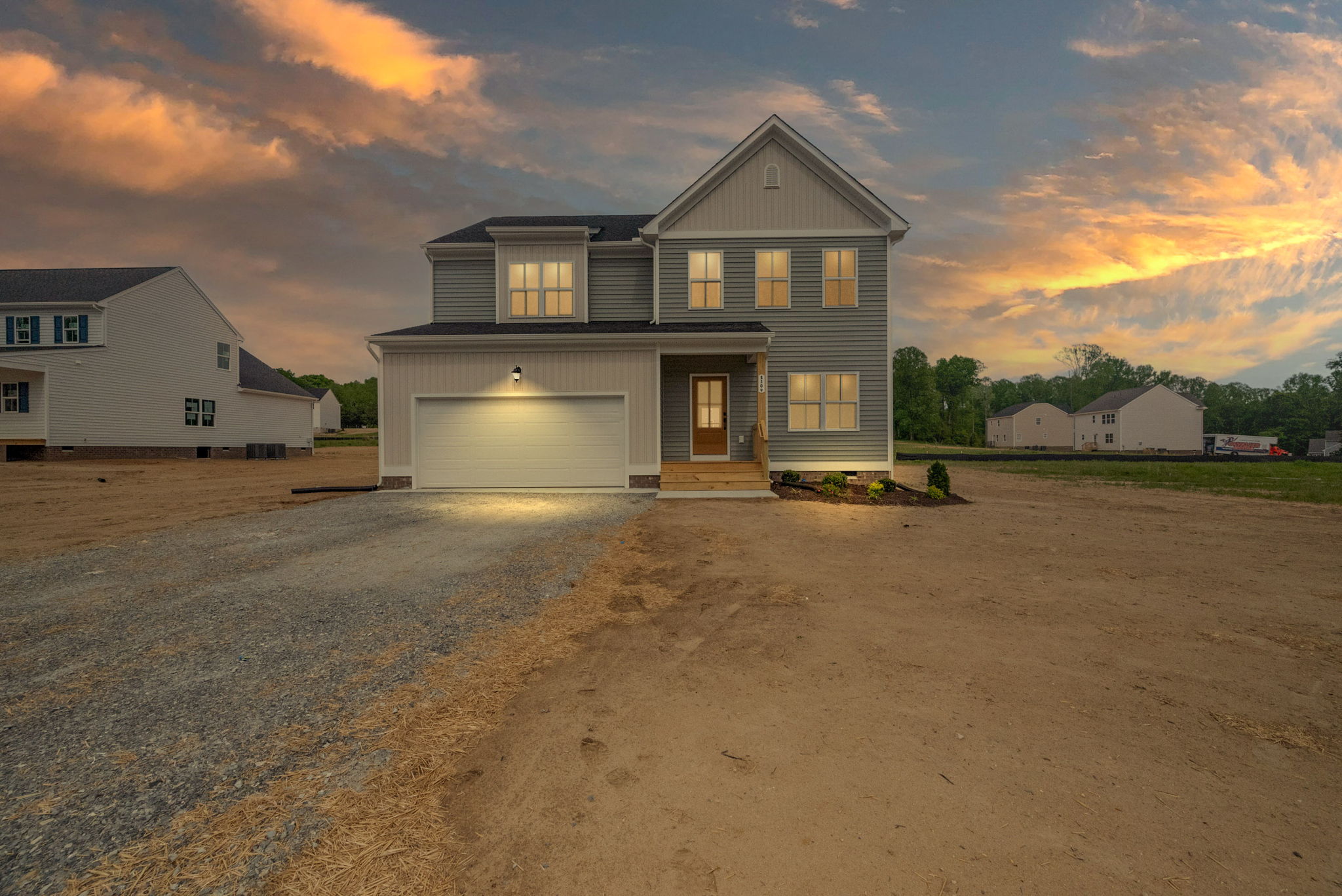 A two-story house with an illuminated garage and front porch, set against a sunset sky. The surroundings are mostly bare ground with sparse greenery. Adjacent houses are visible on either side.