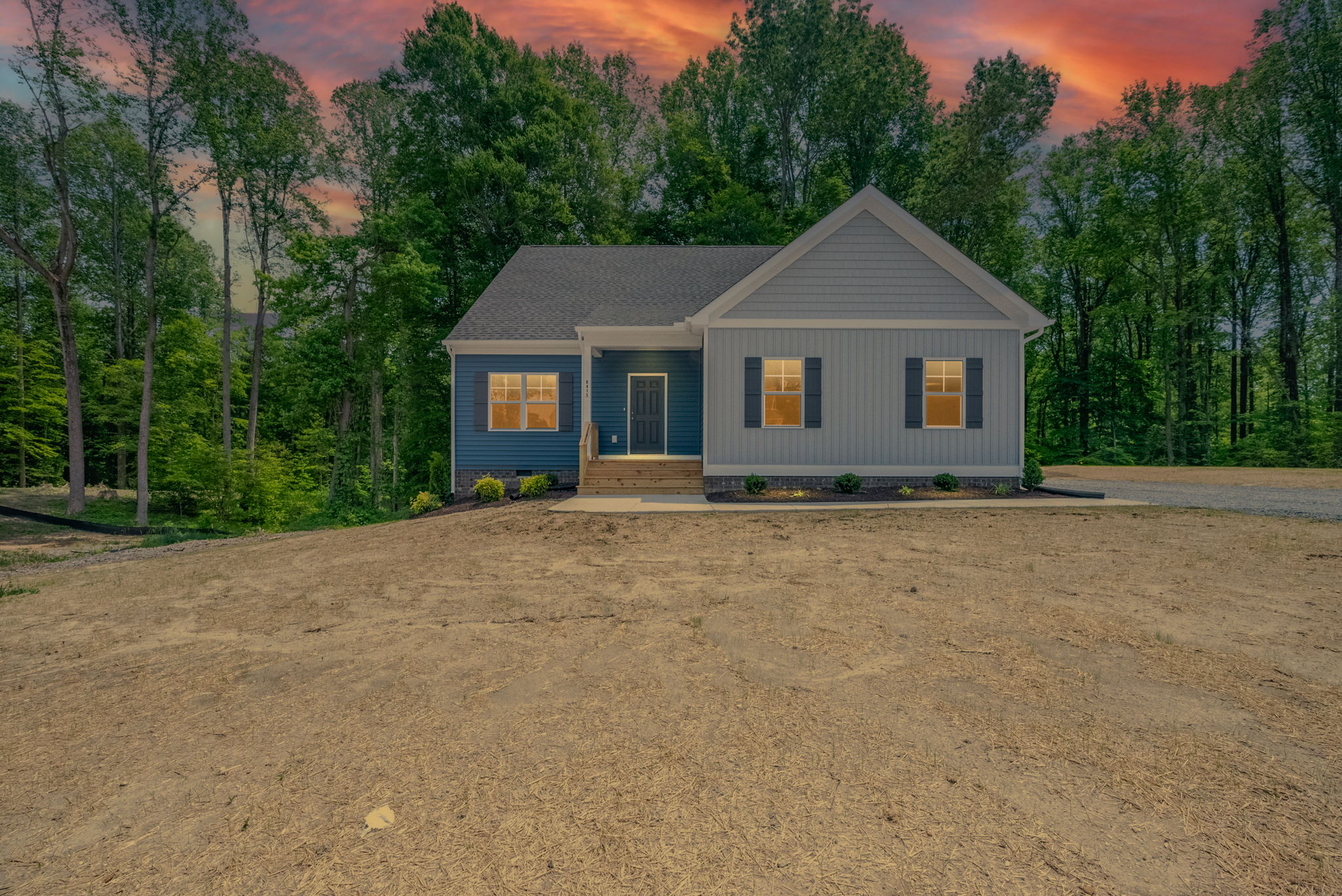 A small, modern single-story house with a gray exterior and white trim, situated on a cleared plot with a backdrop of trees under a colorful sunset sky.