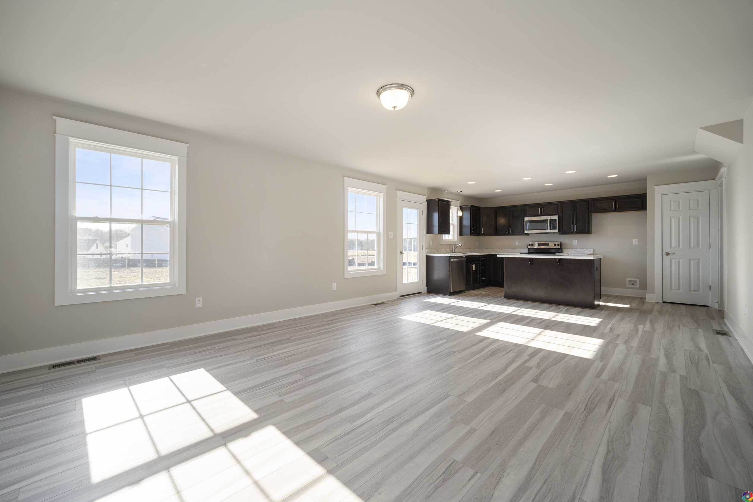 A spacious, unfurnished room with light wood flooring, large windows, and an adjacent modern kitchen featuring dark cabinetry and stainless steel appliances.