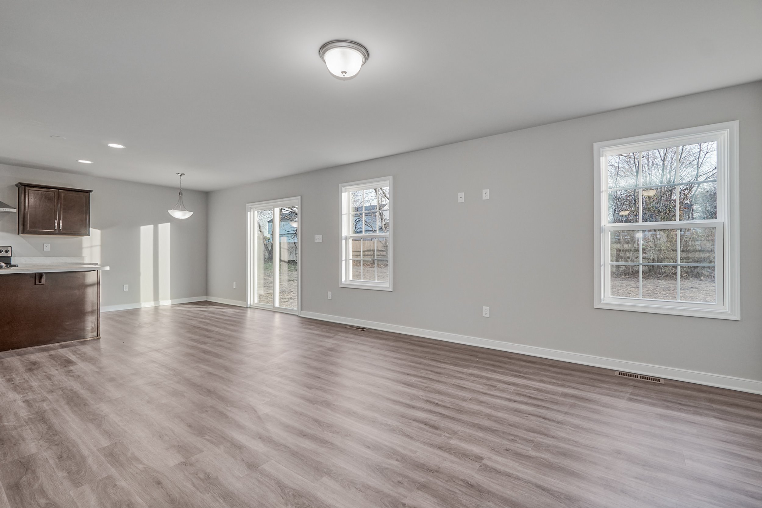 A spacious, unfurnished open-plan living area with light grey walls and wooden flooring, featuring ample natural light from large windows and sliding glass doors leading to an outdoor space.