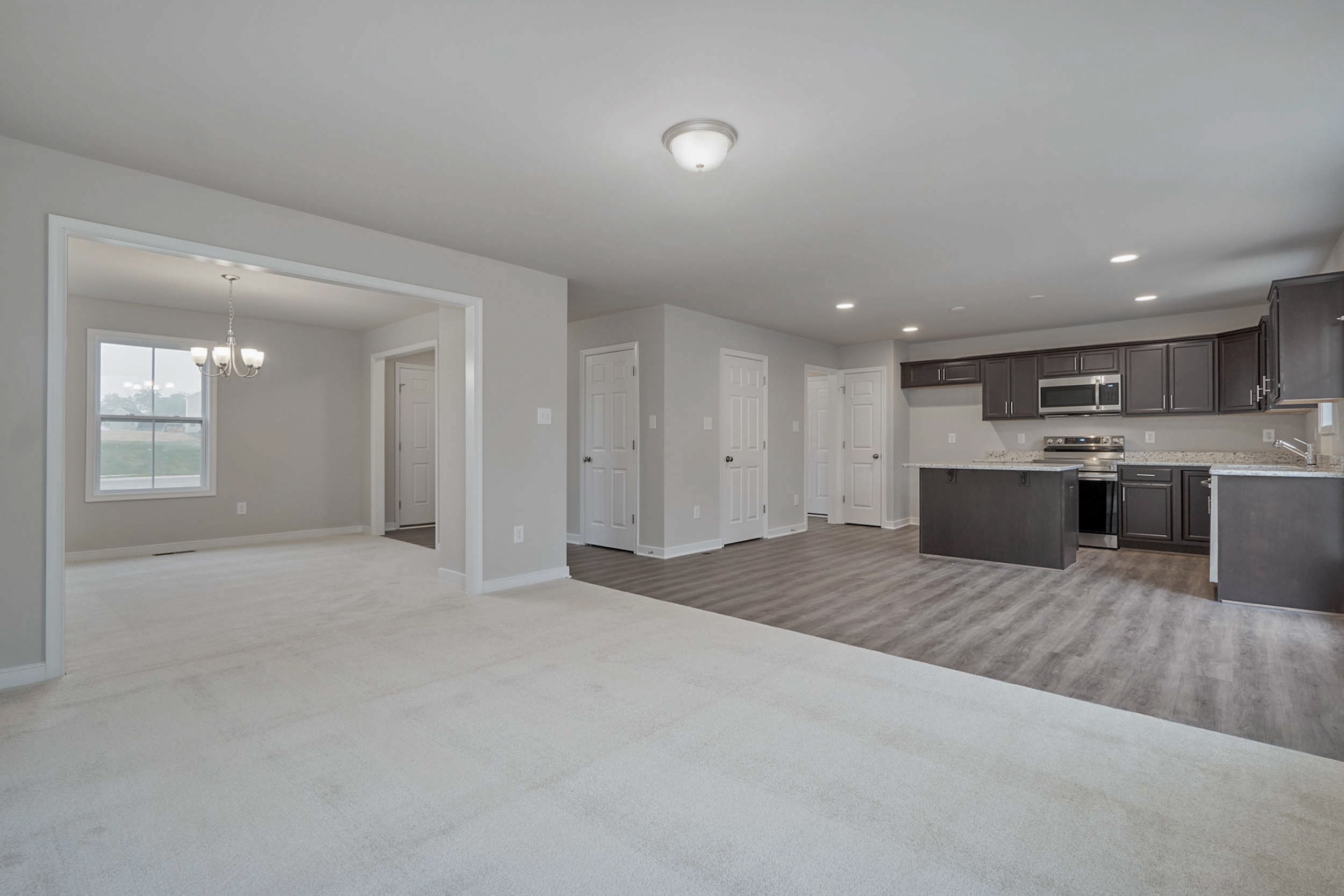 Open-concept living area with carpet, adjacent dining room featuring a chandelier, and a kitchen with dark cabinets, granite countertops, and stainless steel appliances.