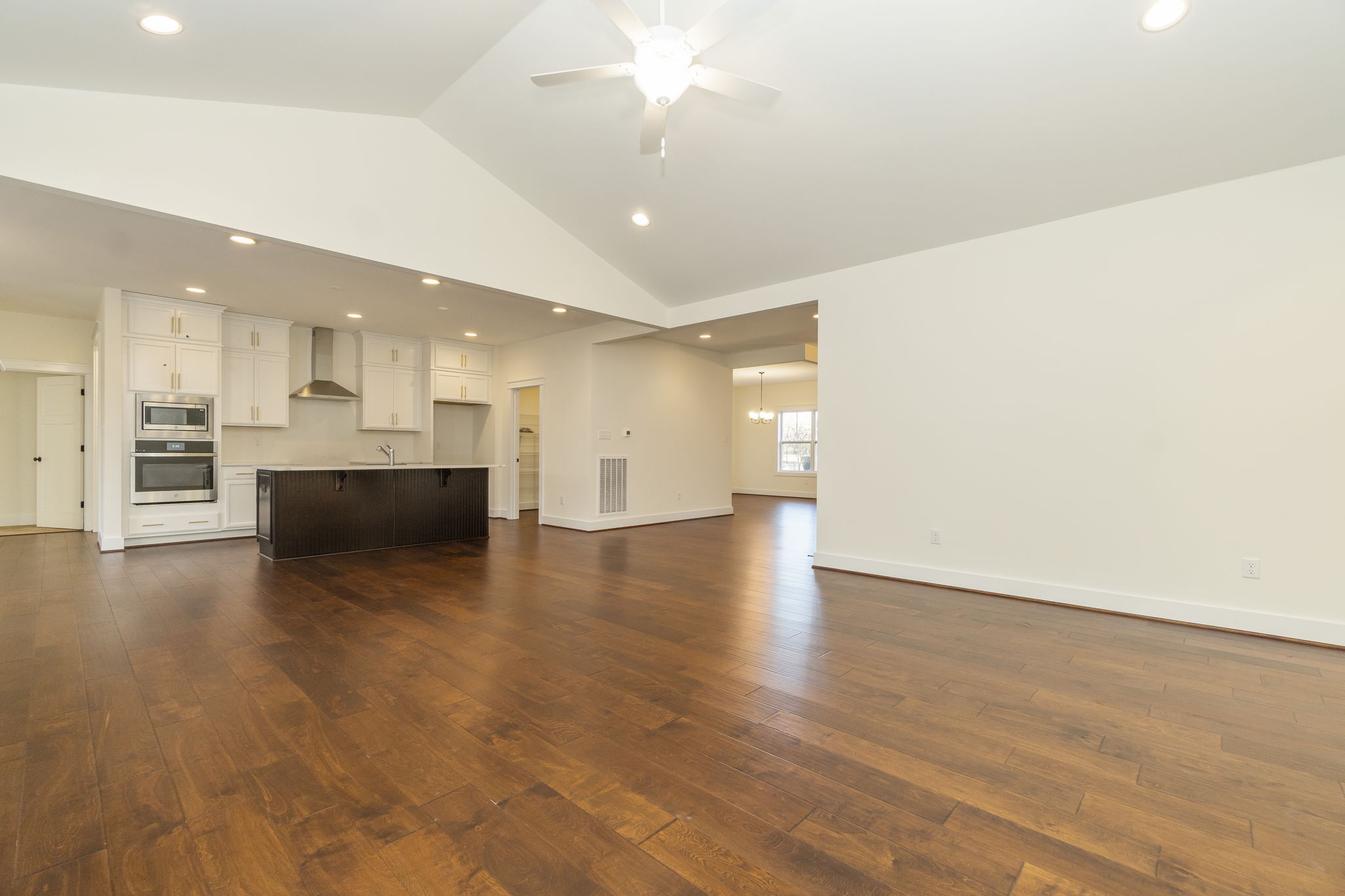 Spacious, empty interior of a modern home featuring hardwood floors, white cabinetry, stainless steel appliances, and vaulted ceilings with a ceiling fan.