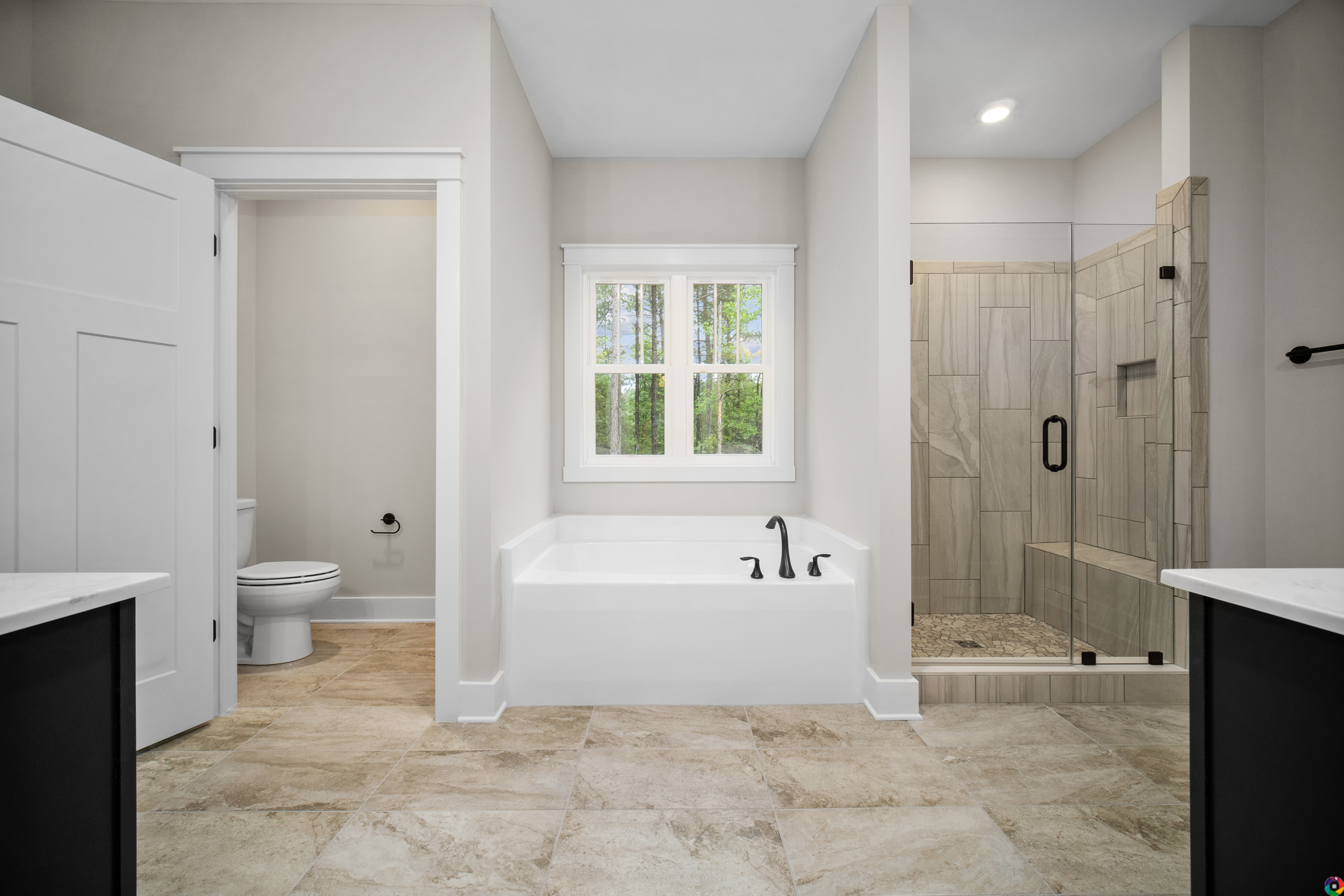 A modern bathroom with a bathtub, shower with glass door, toilet, and double sink vanity. The room has beige tiles and a window above the bathtub.