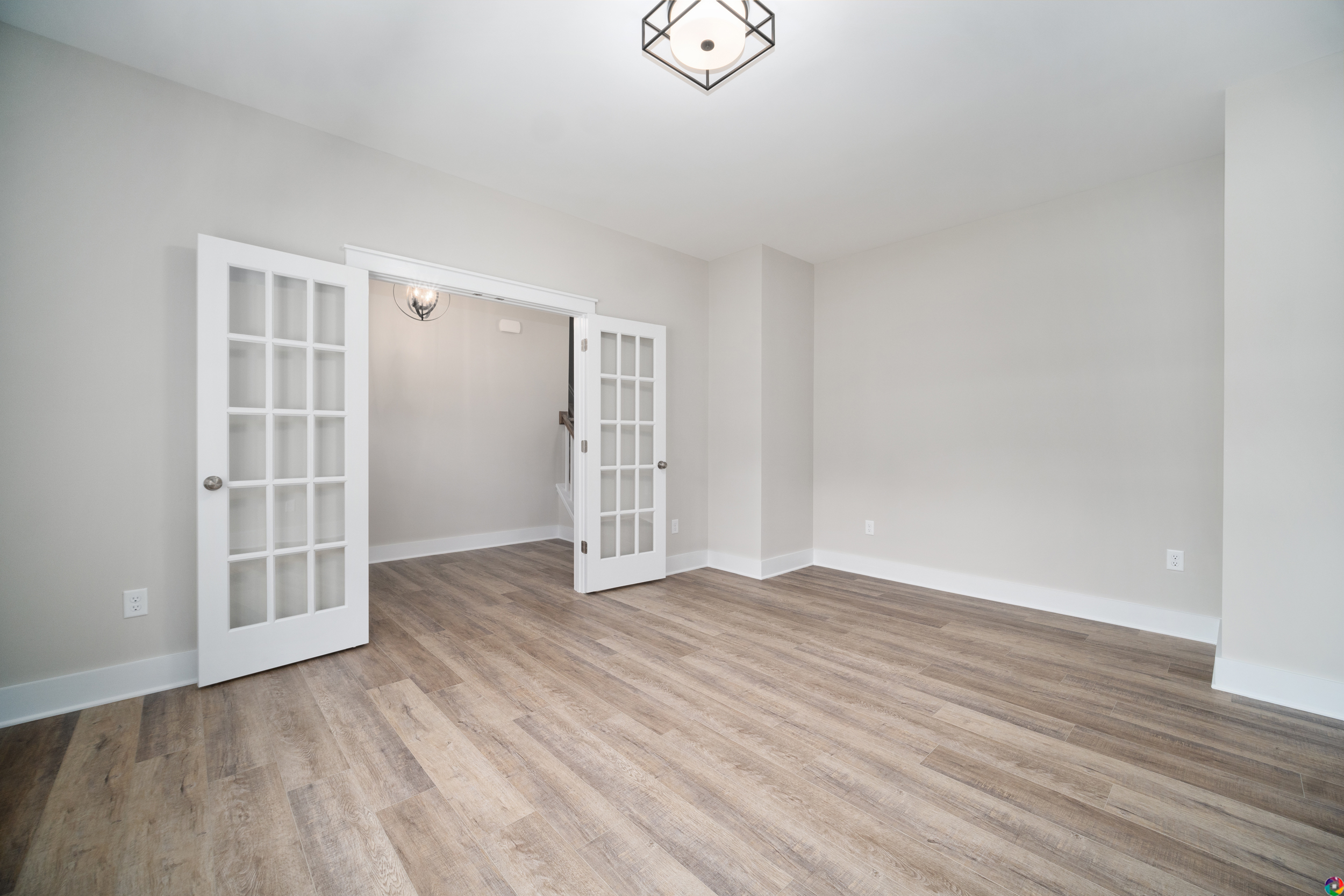 A vacant room with light-colored walls, wooden flooring, and French doors leading to an adjacent space. A modern light fixture is installed on the ceiling.