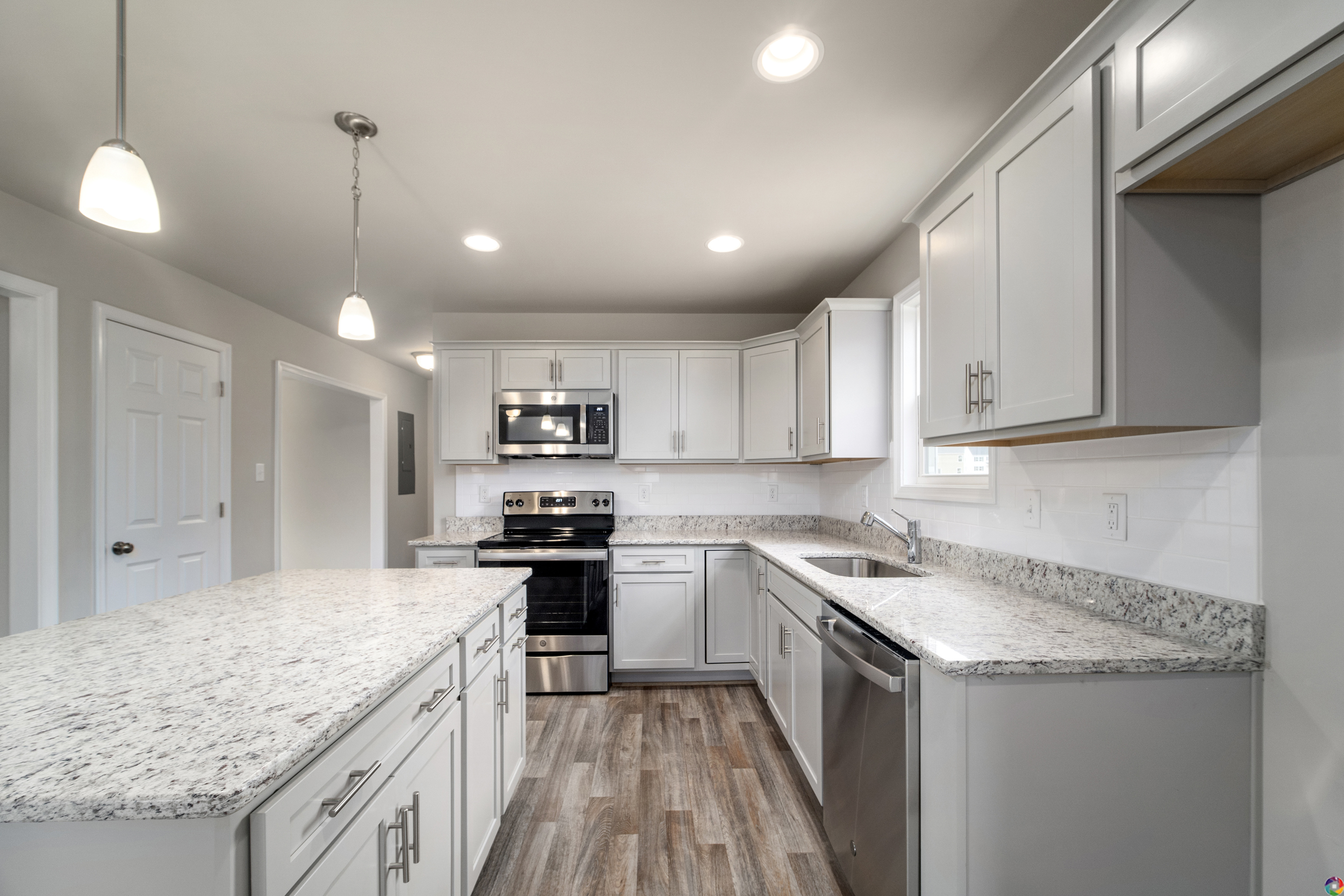 A modern kitchen with white cabinets, granite countertops, stainless steel appliances, a central island, pendant lights, and wood flooring.