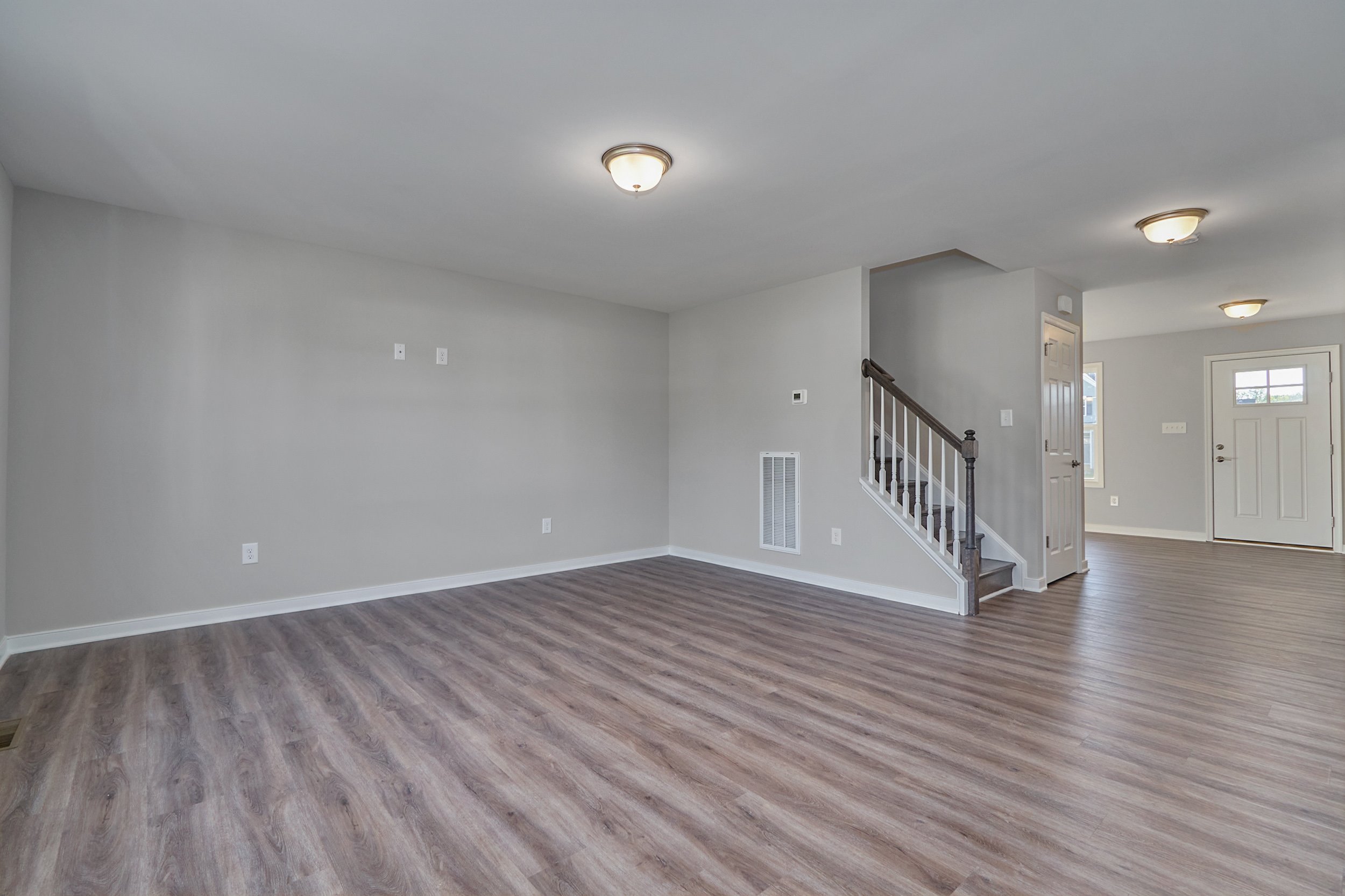 An empty room with wooden flooring, white walls, ceiling lights, and a staircase leading upstairs near a white door with a small window.