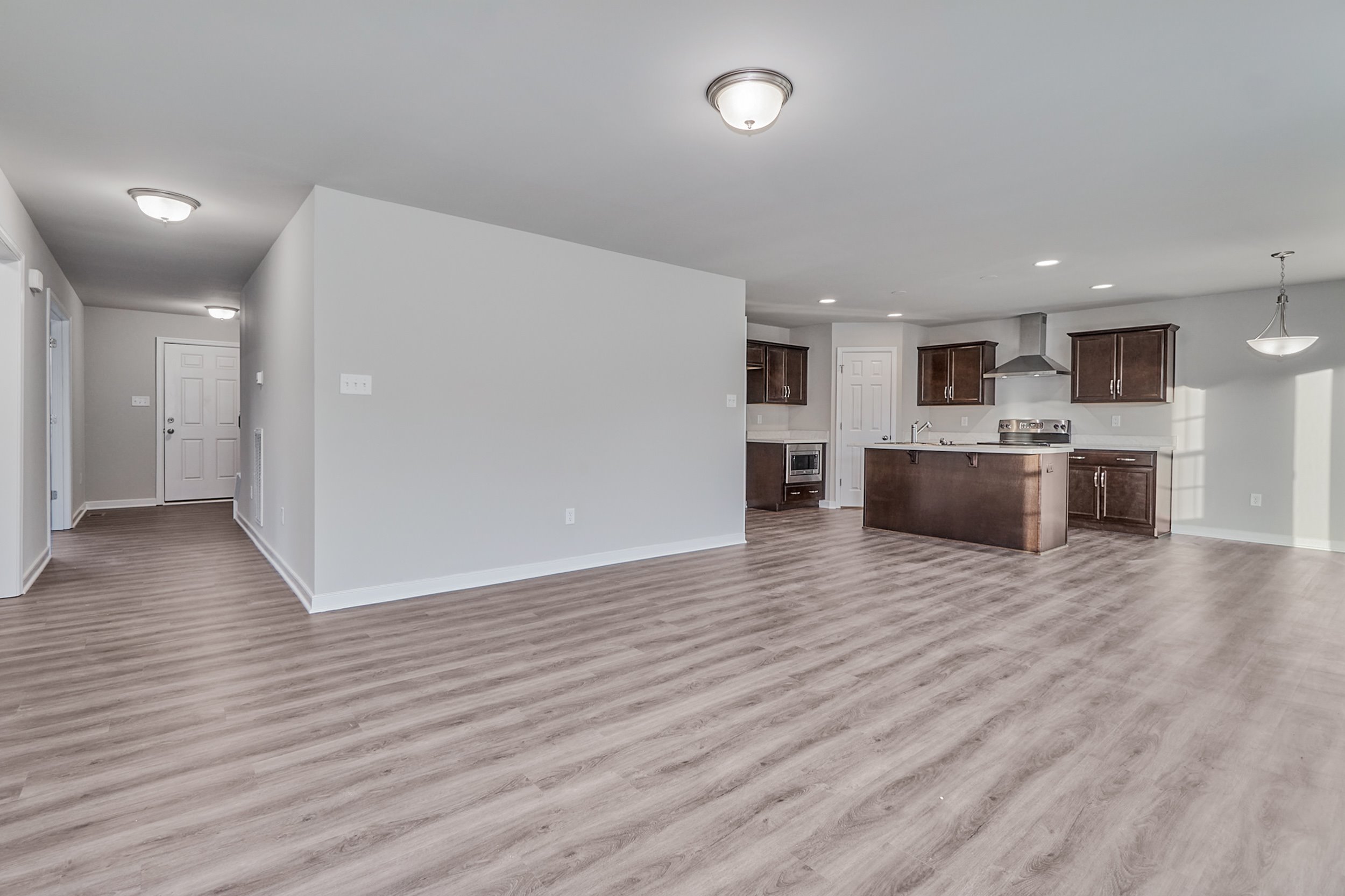 Spacious, empty open-plan living area with light gray walls, wood-look flooring, and a kitchen featuring dark brown cabinets and stainless steel appliances. A hallway leads to a door on the left.