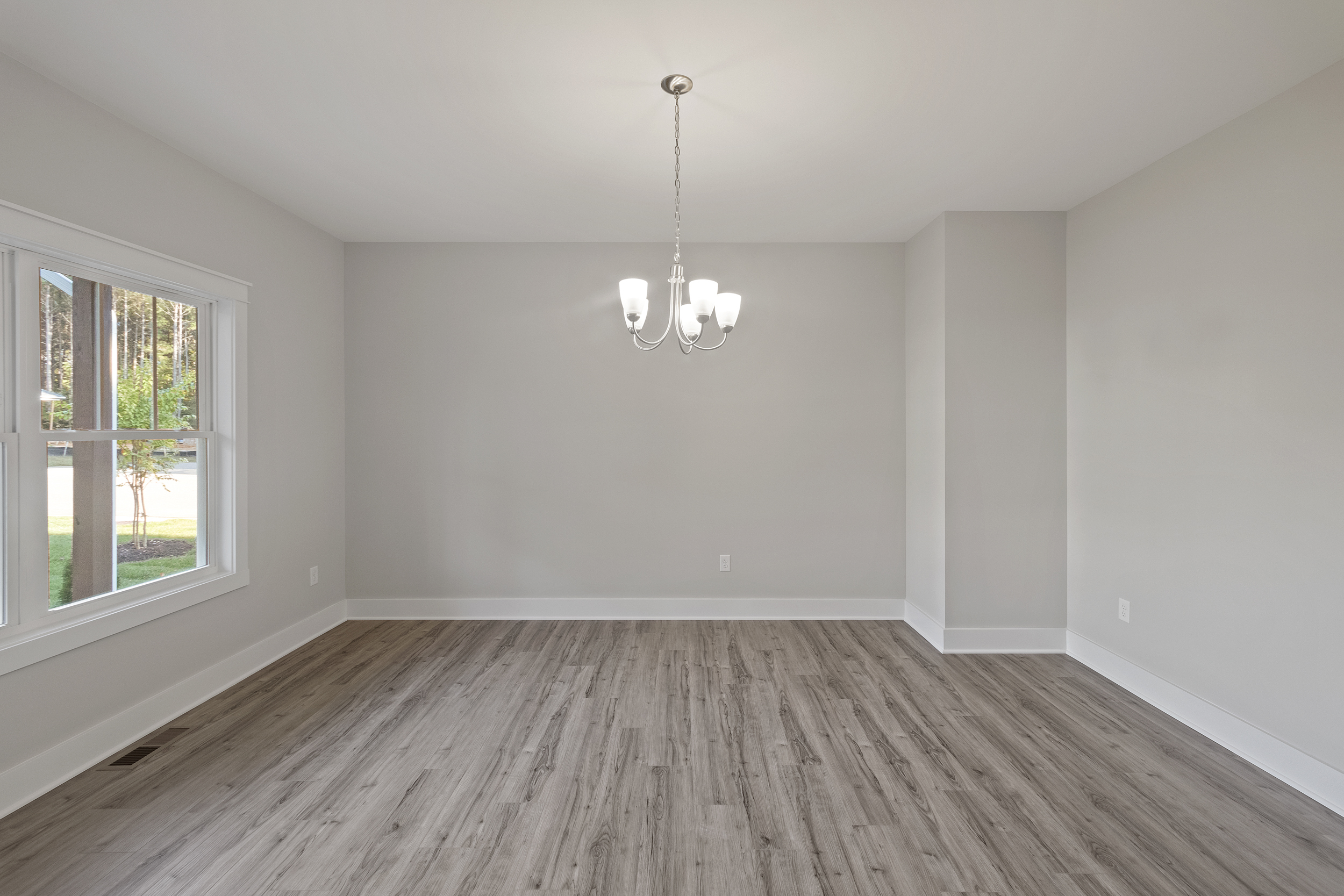 A vacant room with light gray walls, a window, a hanging light fixture, and hardwood flooring.