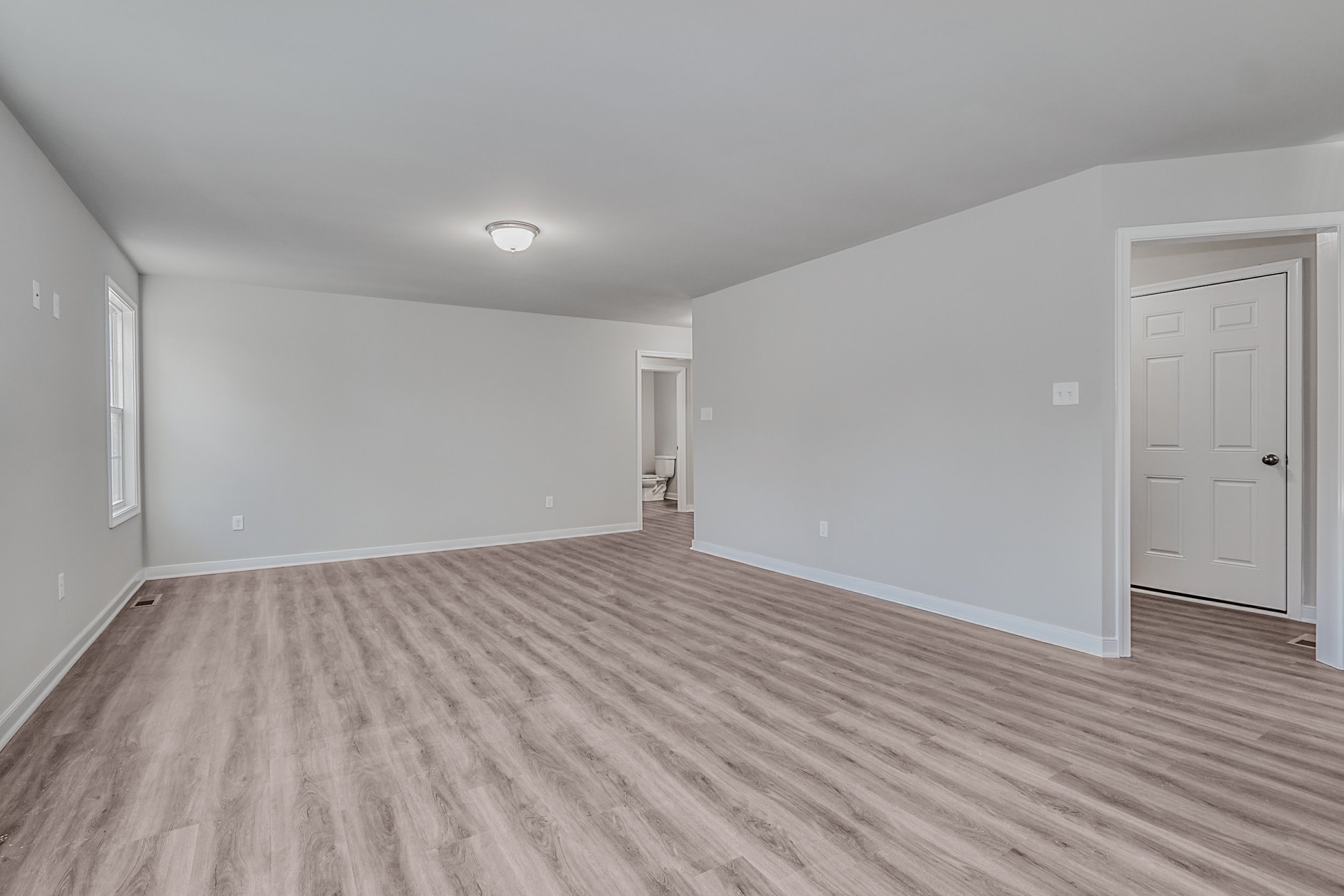 A spacious, empty room with light wood flooring, grey walls, a ceiling light, a window on the left, and a doorway leading to another room.