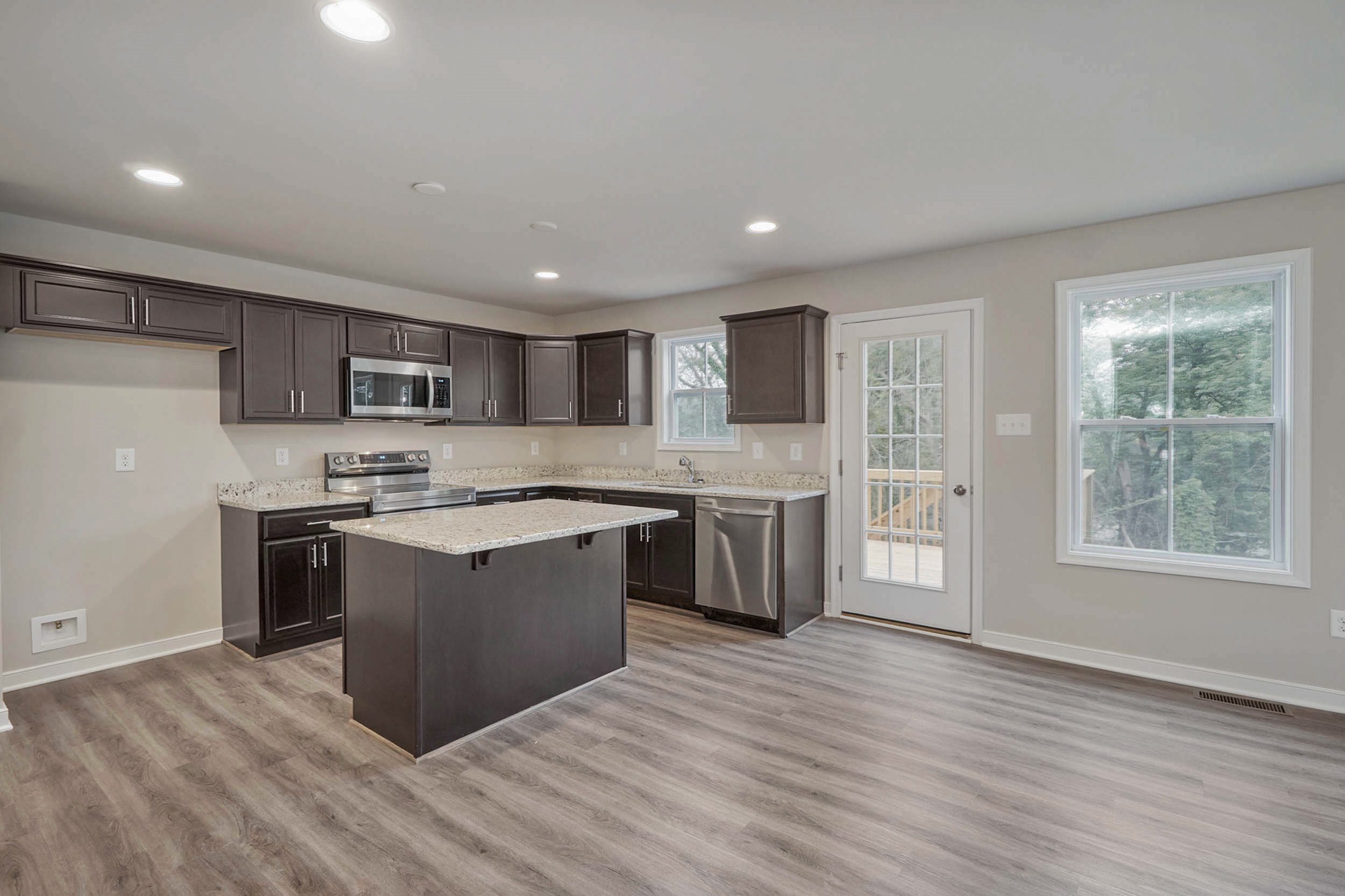 Modern kitchen with dark wood cabinets, stainless steel appliances, and a granite countertop island. The room has hardwood floors, recessed lighting, and a door leading to an outdoor area.
