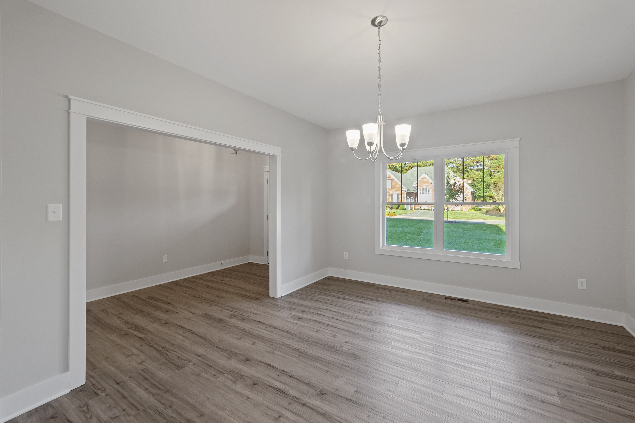 An empty room with light gray walls, a large window, hardwood flooring, and a white-framed chandelier. Another empty space with similar flooring is visible through a wide doorway.