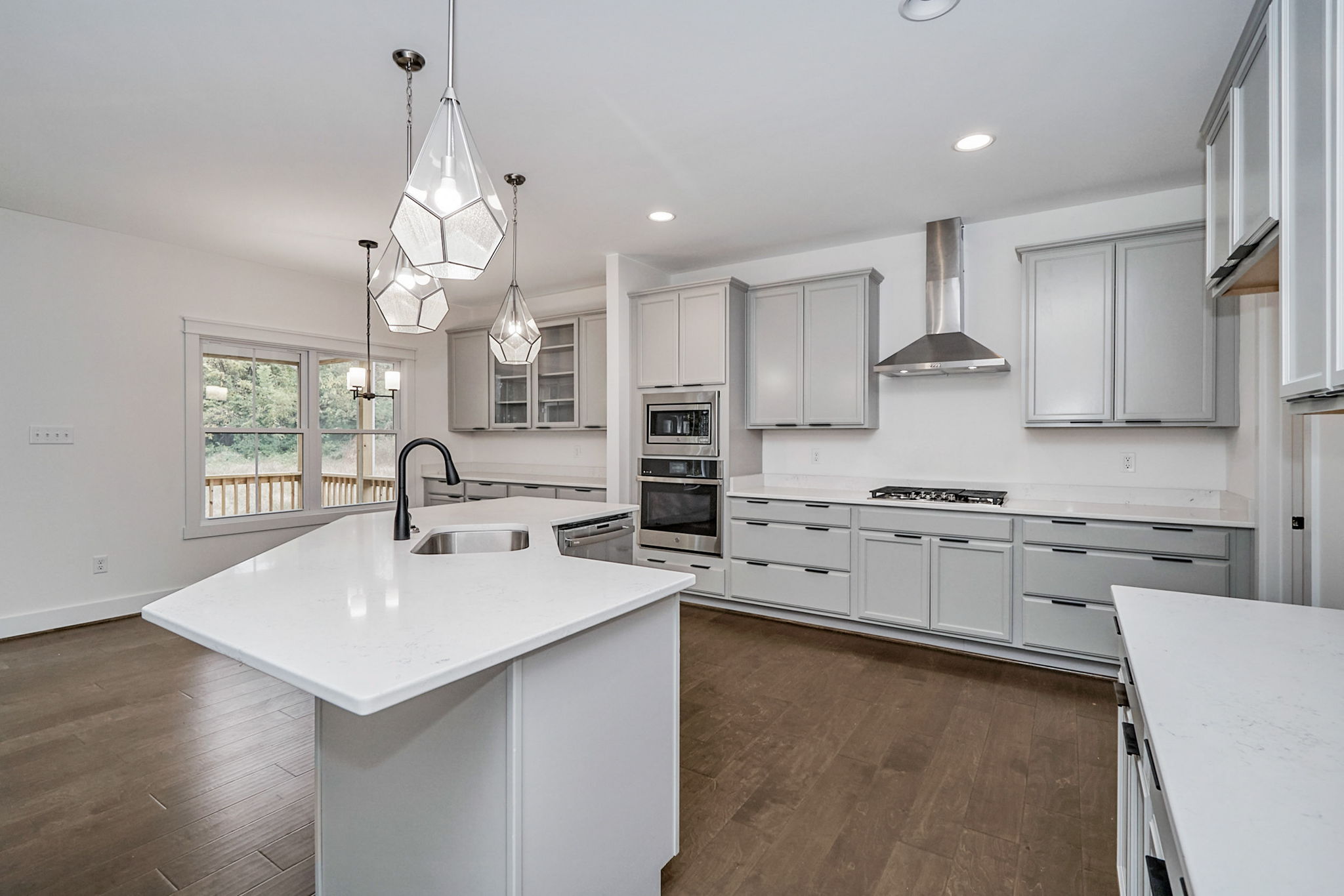 A modern kitchen features light gray cabinetry, a large island with a sink, stainless steel appliances, geometric pendant lights, and hardwood floors, with a view of an outdoor deck through the windows.