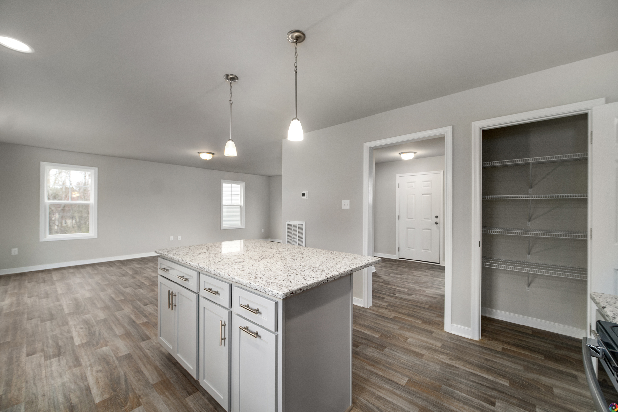 A modern kitchen with granite countertops, an island with white cabinets, pendant lights, hardwood flooring, an open pantry, and a view of a bright, spacious living area.